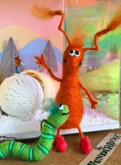 A felt craft beetle with googly eyes and pink feet is waving, while a green plush worm with black stripes stands nearby. The background has whimsical mountains and a miniature skier on a scoop of ice cream snow.
