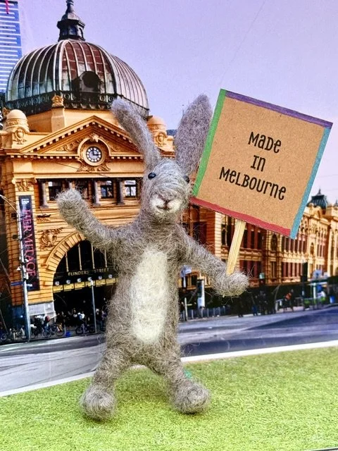 A felt craft rabbit holding a sign that says 'Made in Melbourne', standing on a green surface outside Flinders Street Station in Melbourne, Australia.