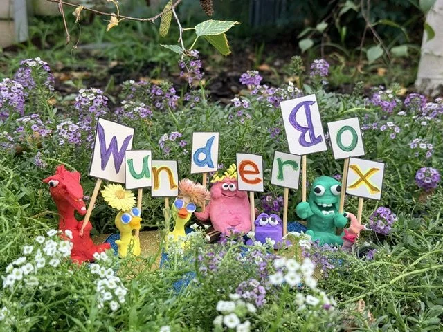 Colorful clay characters holding signs that spell out "Wunderbox" in a garden with purple and white flowers.