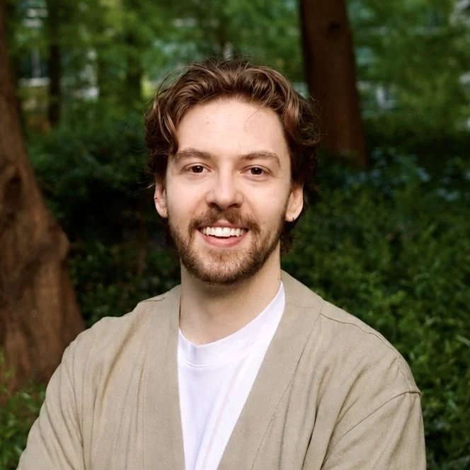 Zac with curly brown hair and a beard, smiling, standing outdoors in a forested area with green foliage in the background.