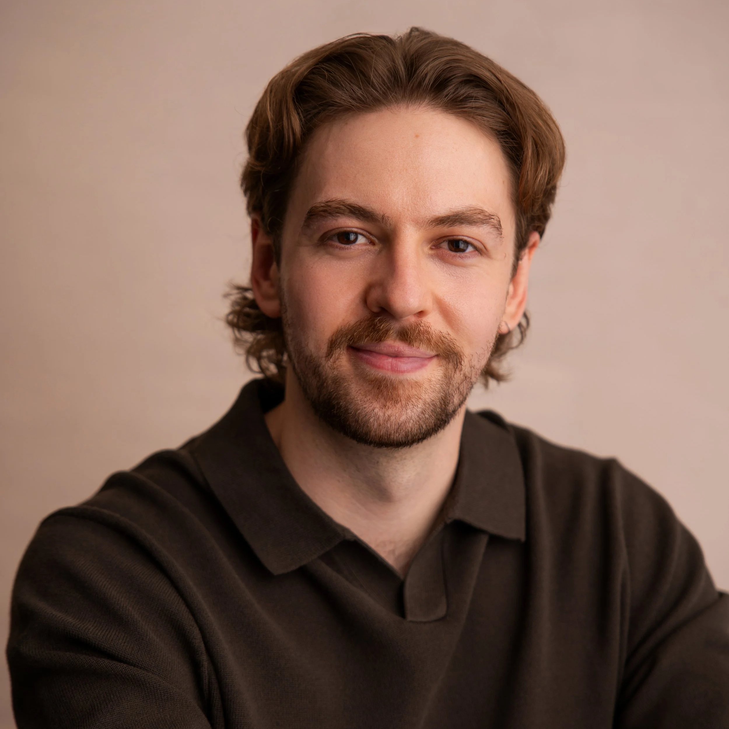 Close-up of Zac, a young man, brown wavy hair, and a beard, smiling slightly, wearing a black shirt, against a plain beige background.