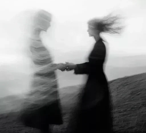 Two women holding hands on a grassy hill, with the woman on the right having hair blowing in the wind, in a black-and-white photo.