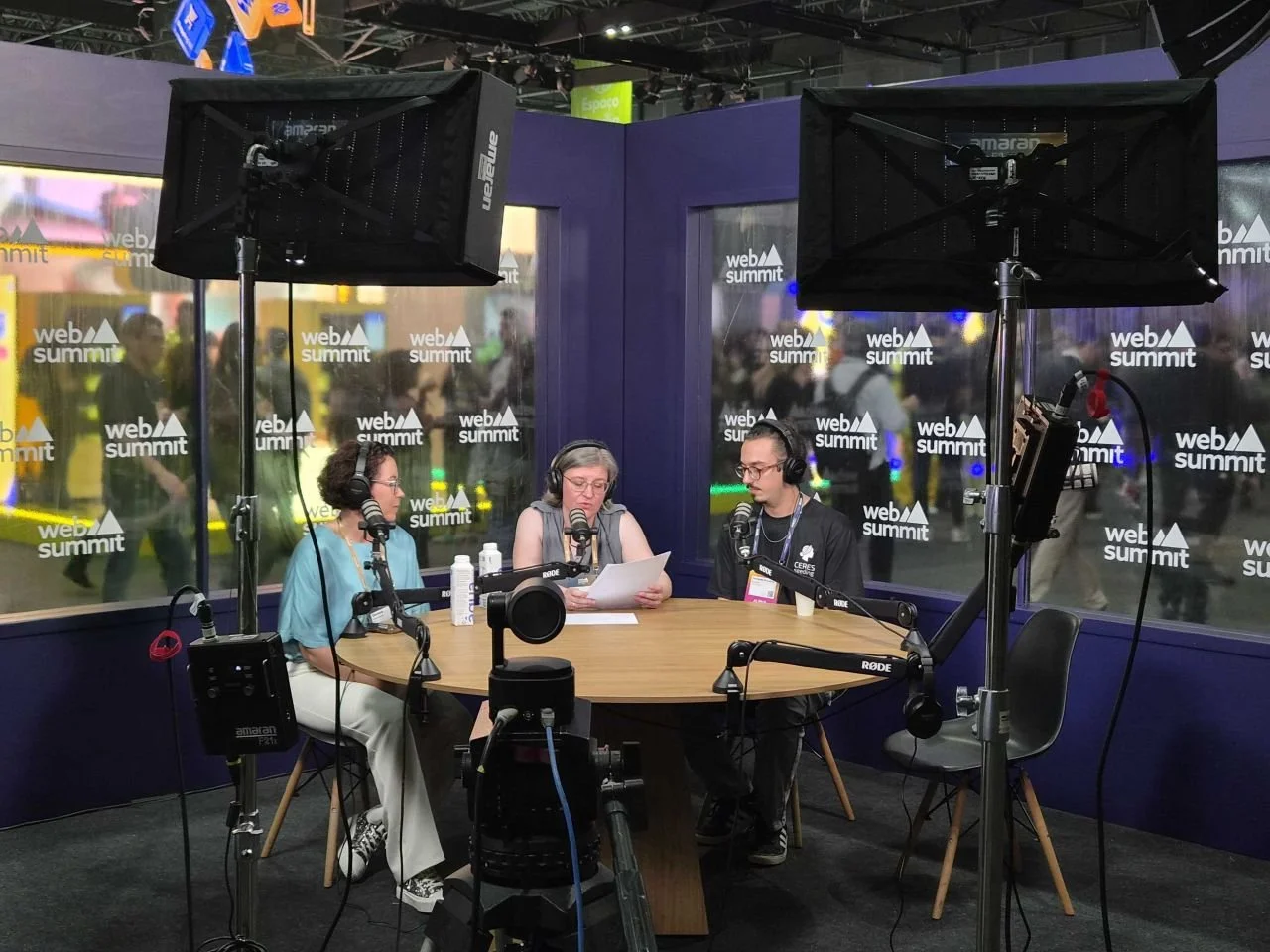 Three people in a podcast recording studio with sound equipment, microphones, and studio lights, with a large window showing a busy convention or event outside.