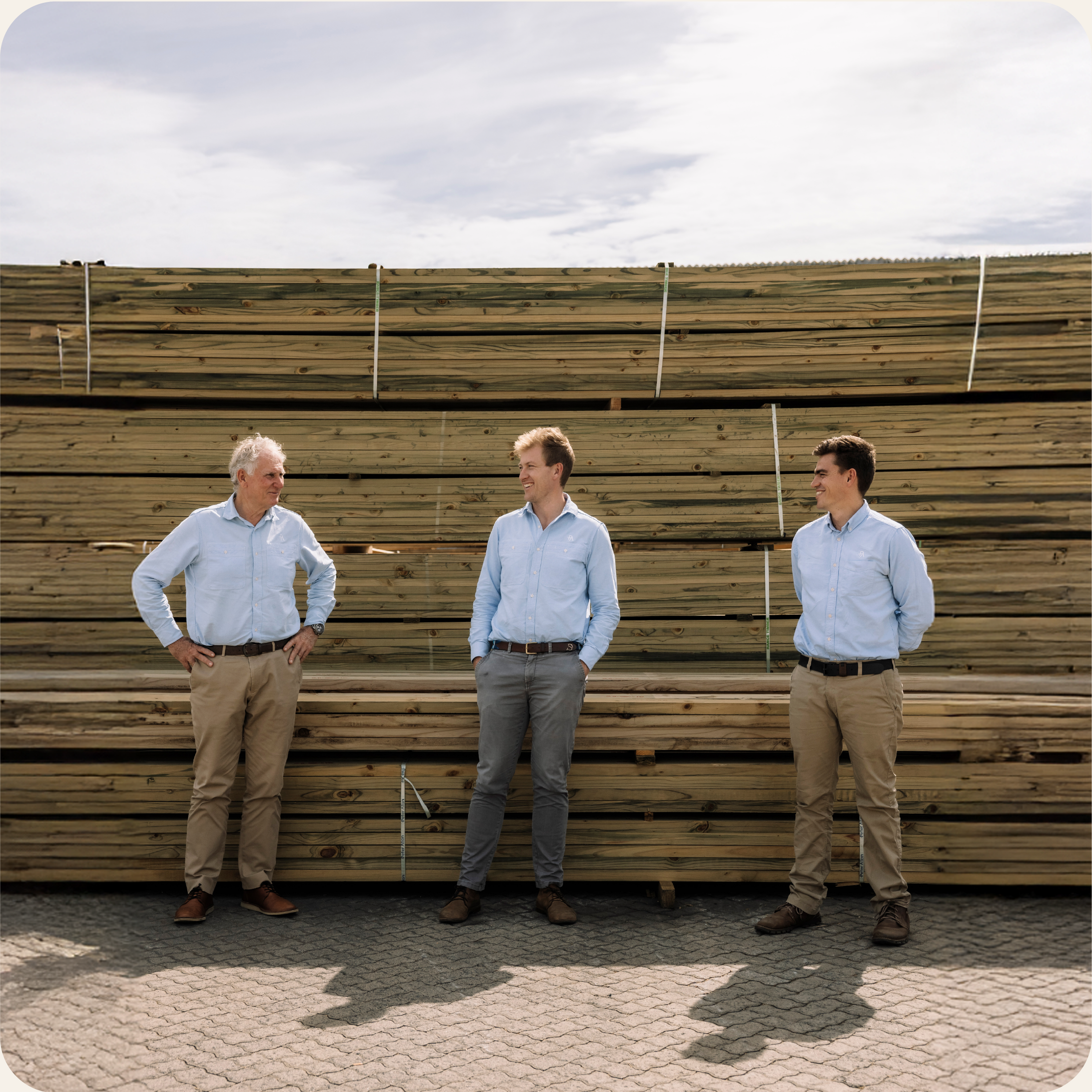 Four men in light blue shirts standing outdoors in front of a large stack of wooden planks, with a cloudy sky overhead.