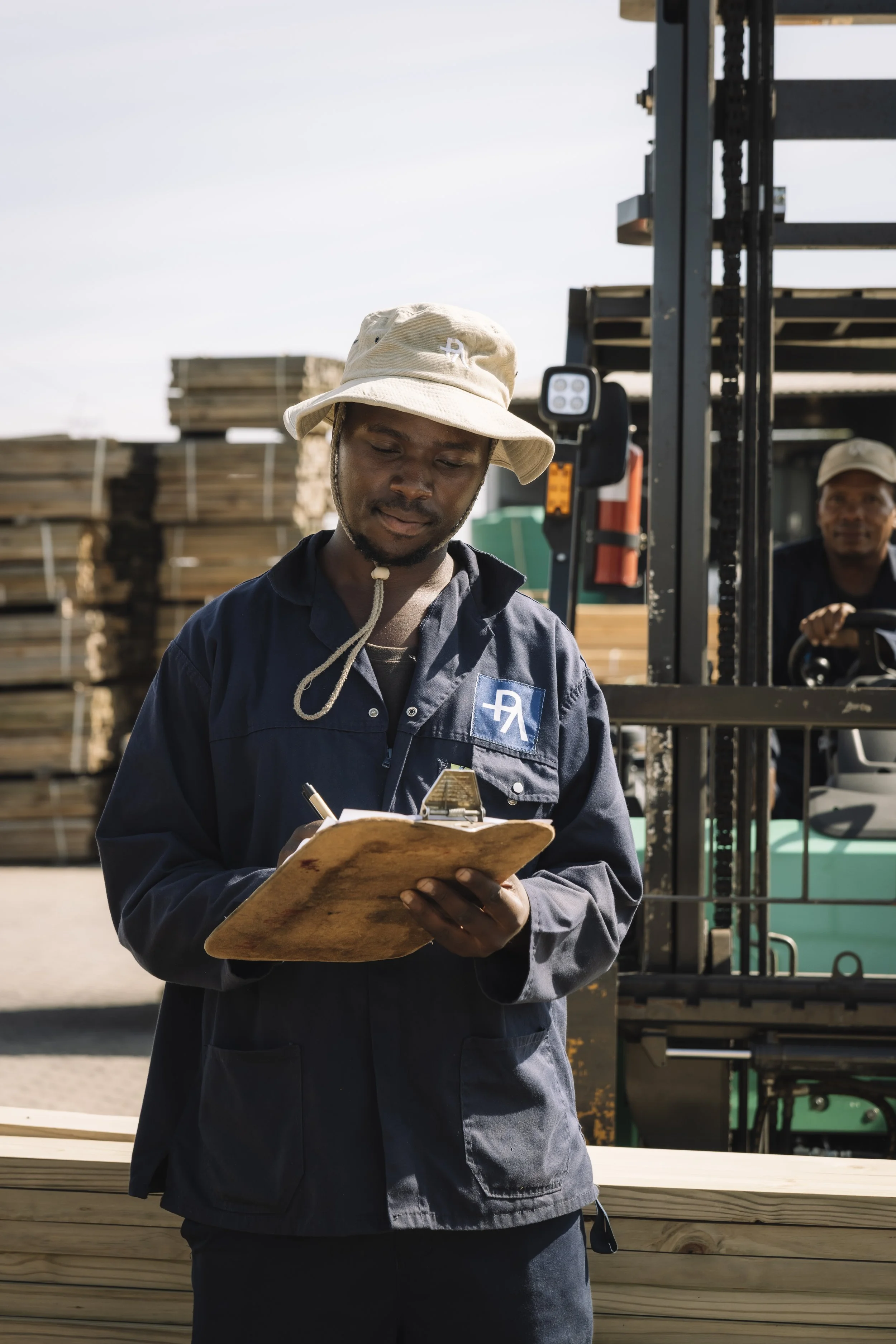 A man in a beige bucket hat and dark work uniform writing on a clipboard outdoors, with a forklift and stacks of lumber in the background. Another man is visible in the background operating the forklift.
