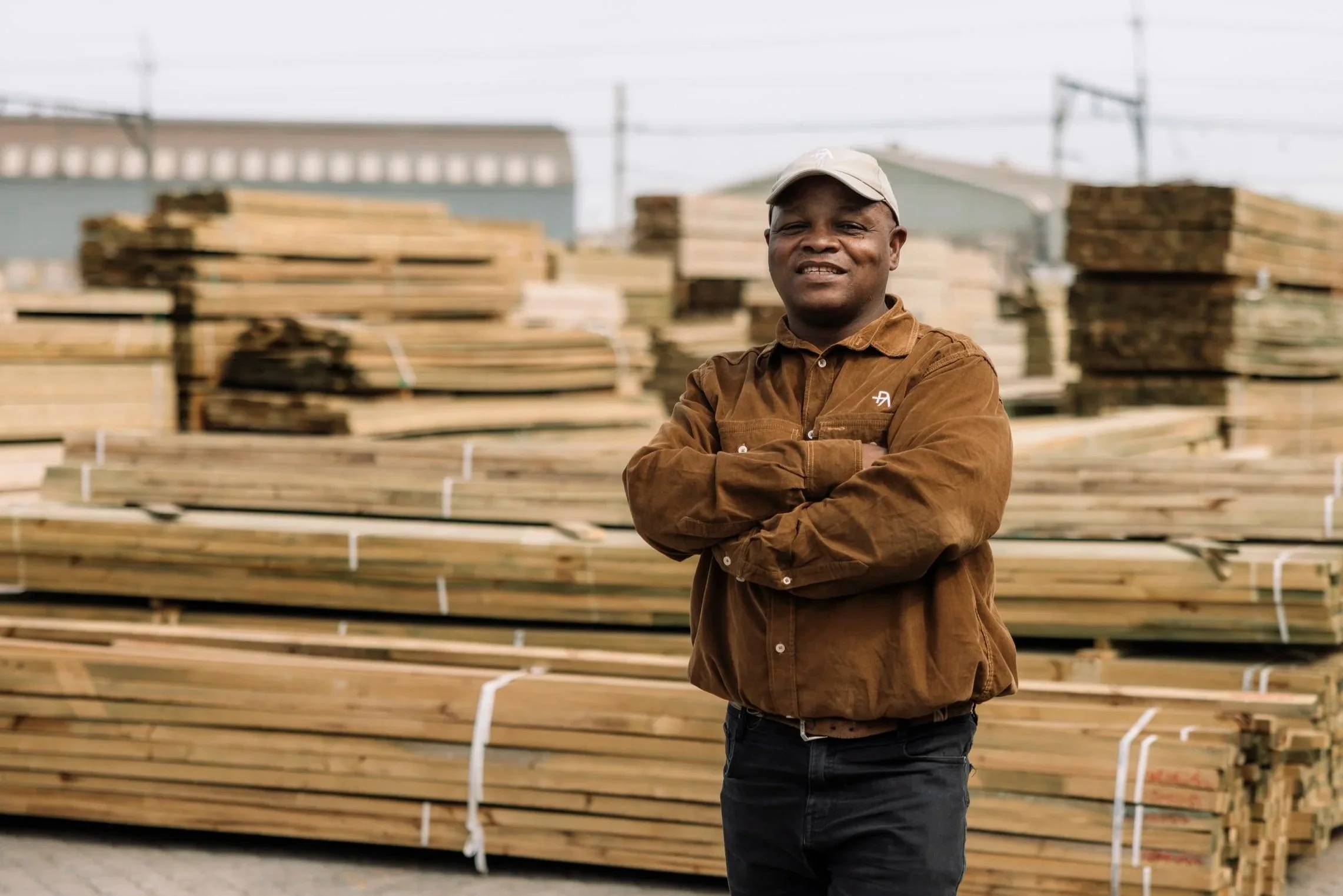 A man with crossed arms wearing a brown jacket and a beige cap standing in front of stacked lumber outdoors.