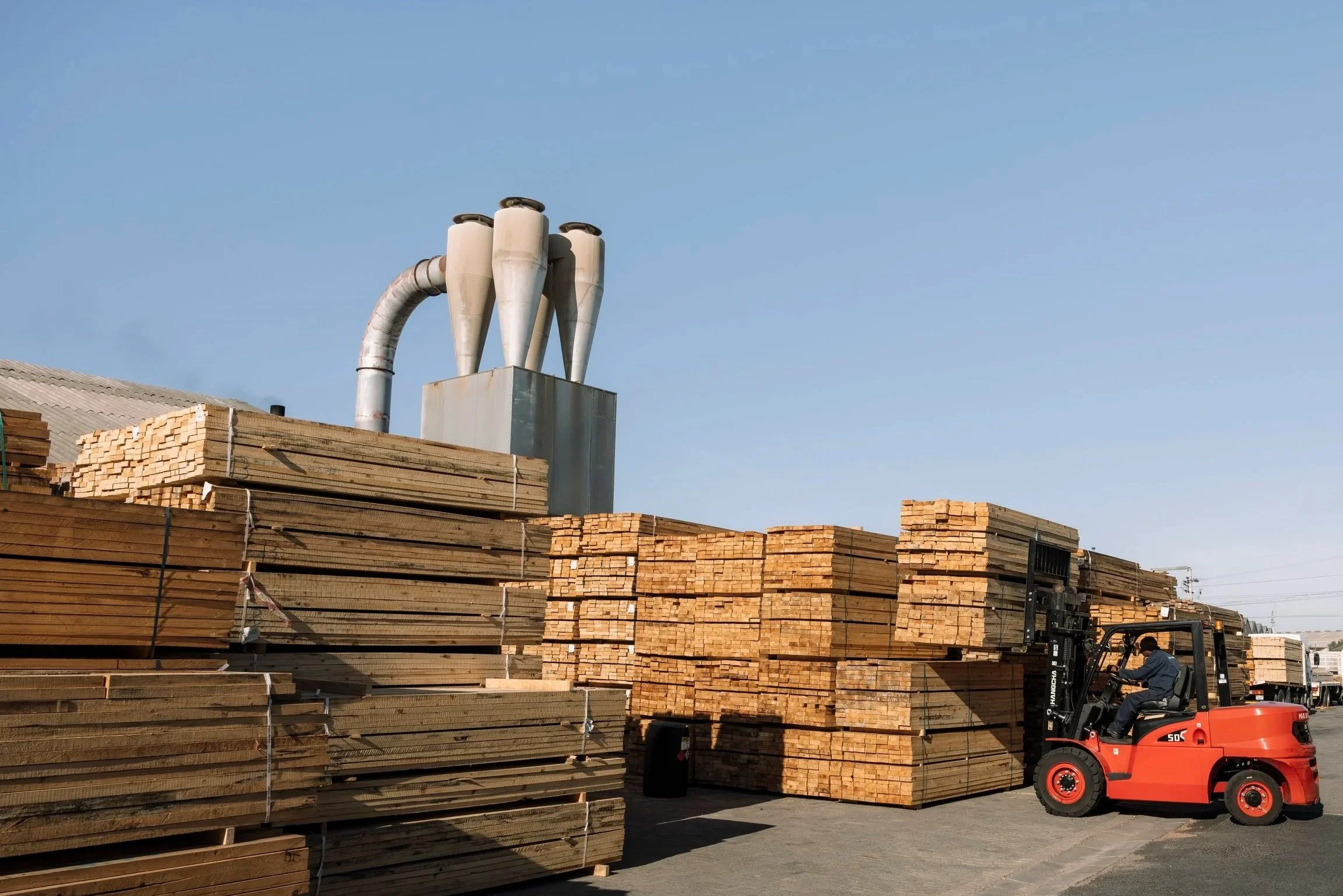 A forklift moving stacks of large wooden planks in a timber yard with a clear blue sky in the background.