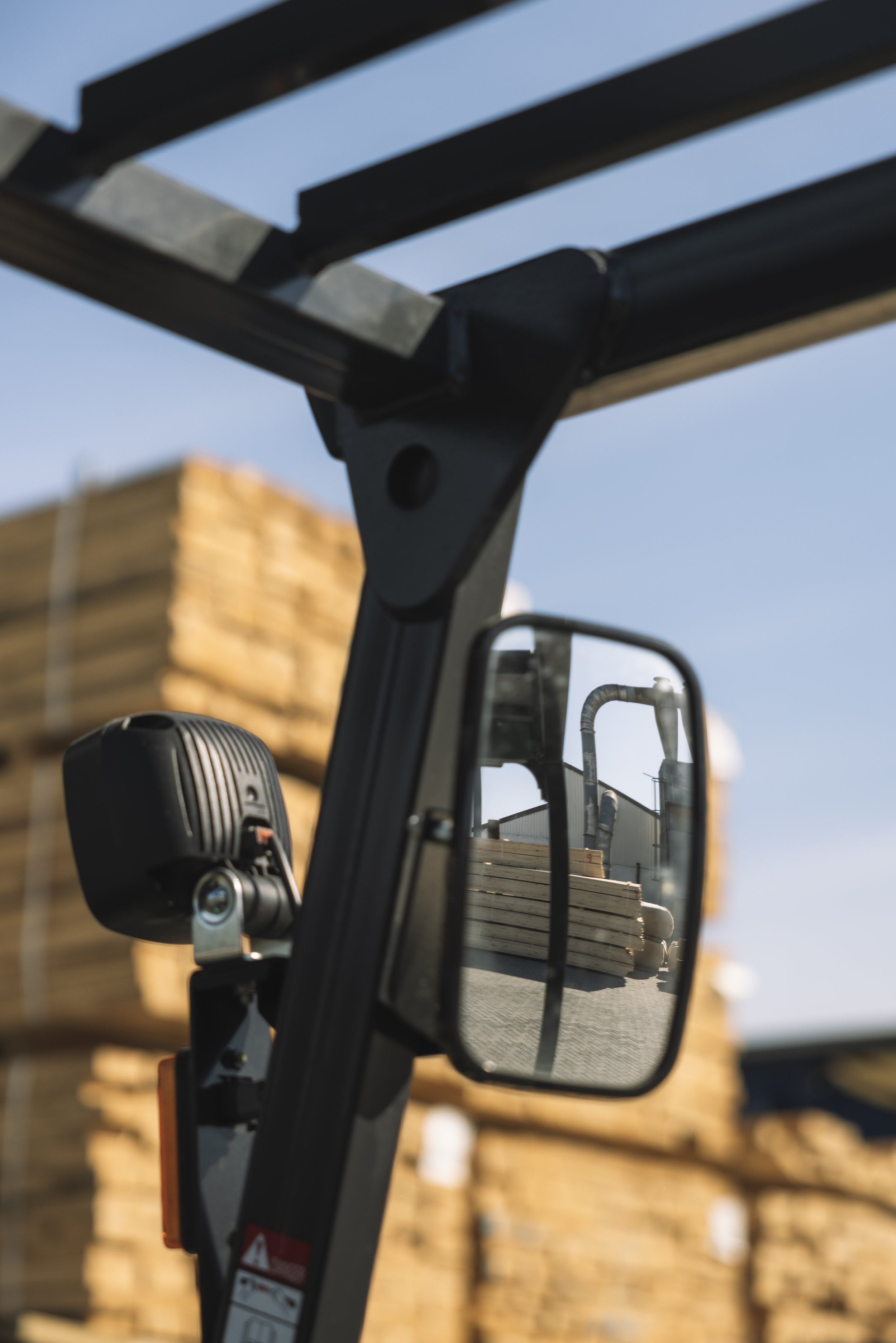 Close-up of a forklift mirror reflecting a stack of lumber, with a blue sky in the background.