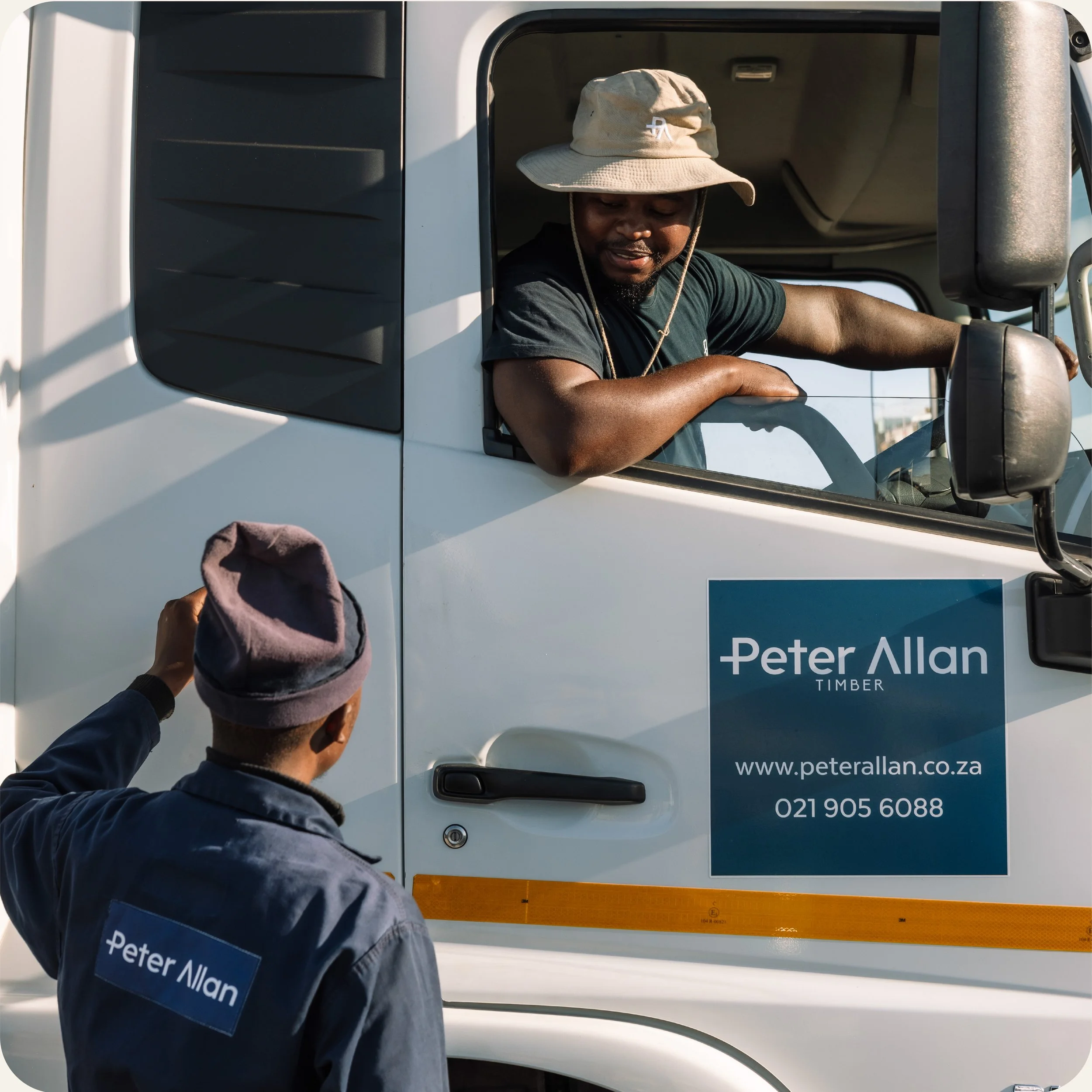 A man wearing a beige wide-brimmed hat and a dark blue jacket with an embroidered name, leaning out of the open window of a white service van. The van has a blue sign with white text that reads "Peter Allan TIMBER" and the website "www.peterallan.co.za." There is also a phone number visible below the sign, and a person in the foreground is holding a cap near the van.