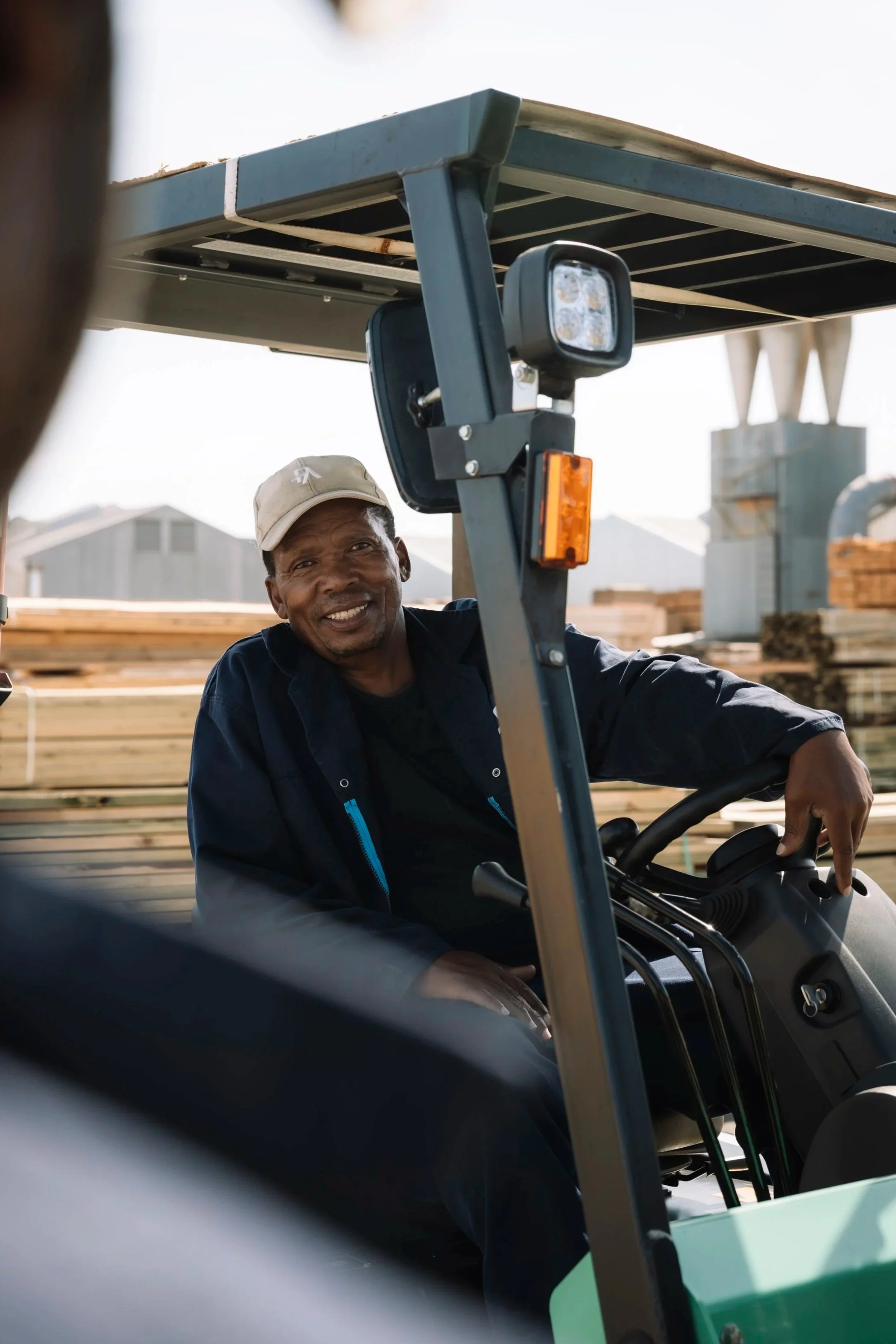 A man smiling while sitting on a green and black forklift, wearing a beige cap and a dark jacket, in an outdoor industrial area with building materials in the background.