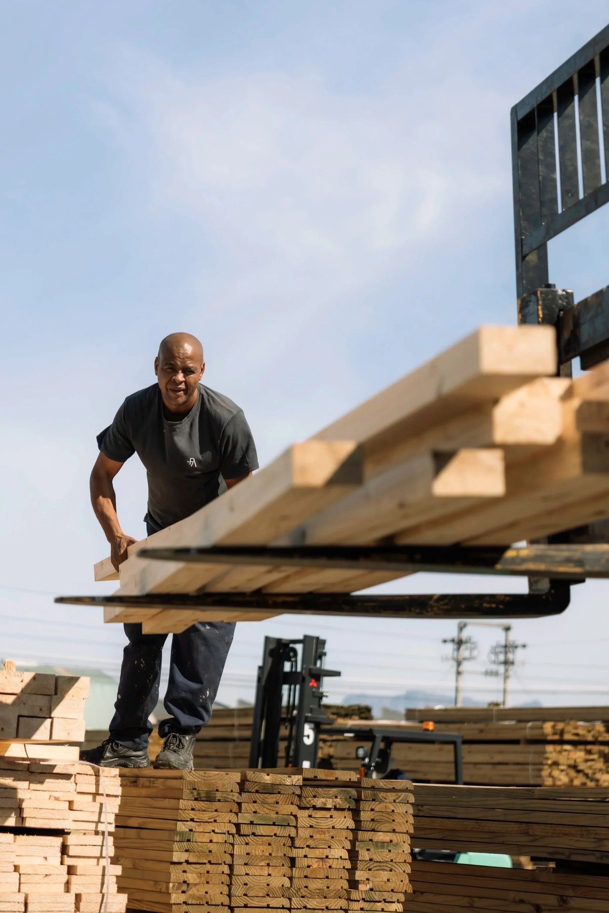 A man working on a construction site lifting wooden planks, with additional stacks of wood and construction equipment visible outside.