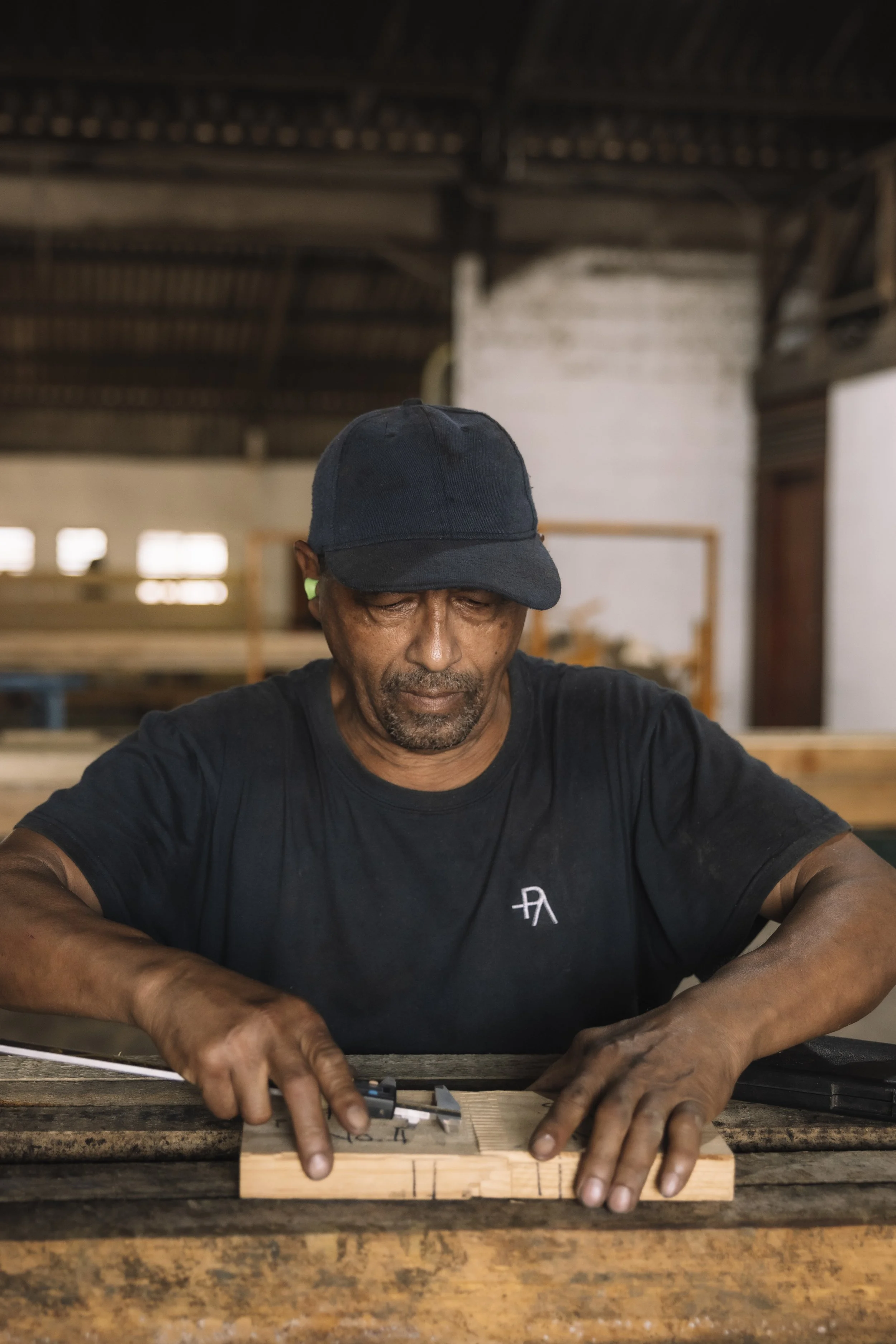 A man in a black shirt and dark cap working on a piece of wood at a workshop.