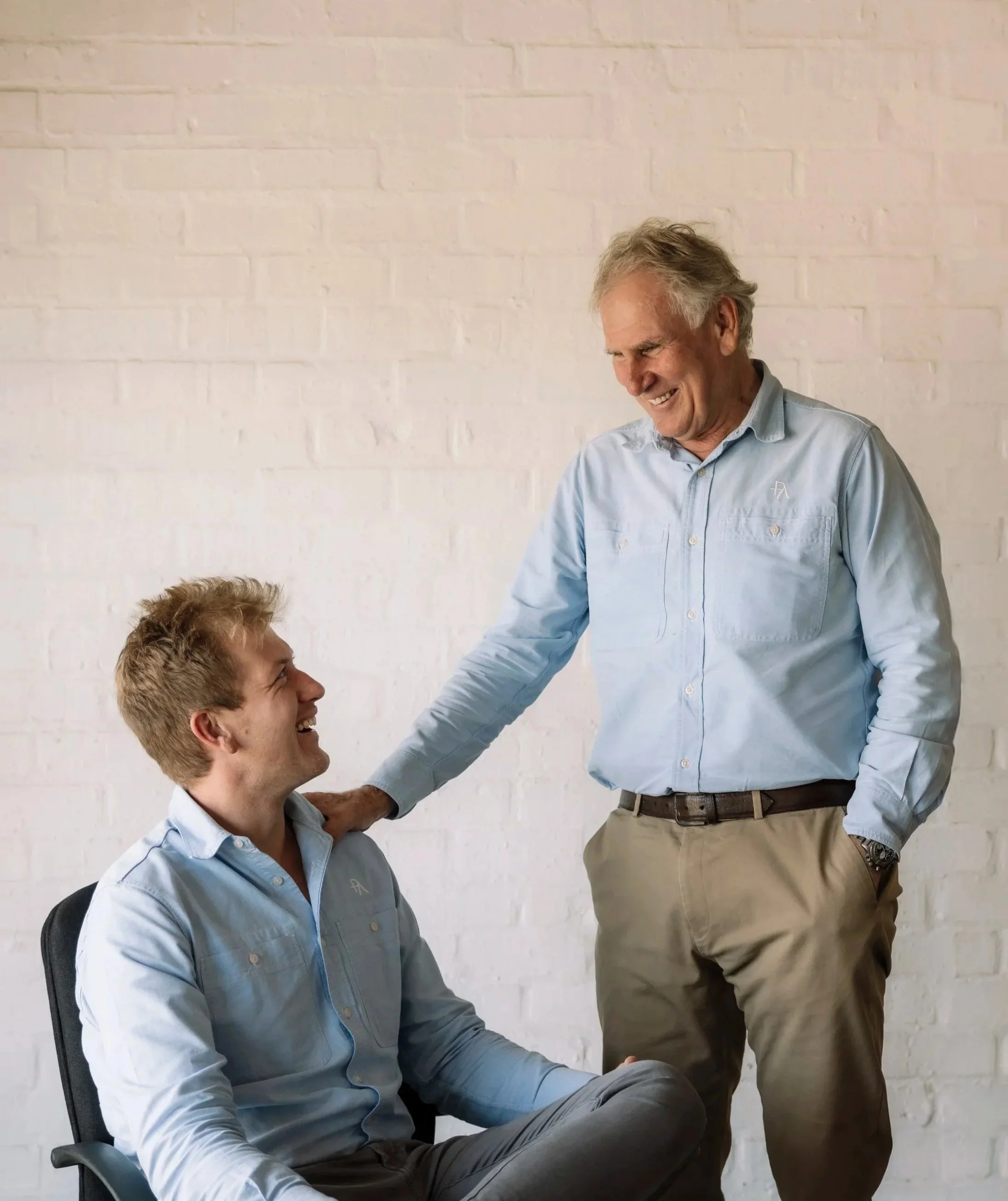 Two men, one seated and the other standing, engaging in conversation and smiling, against a white brick wall.