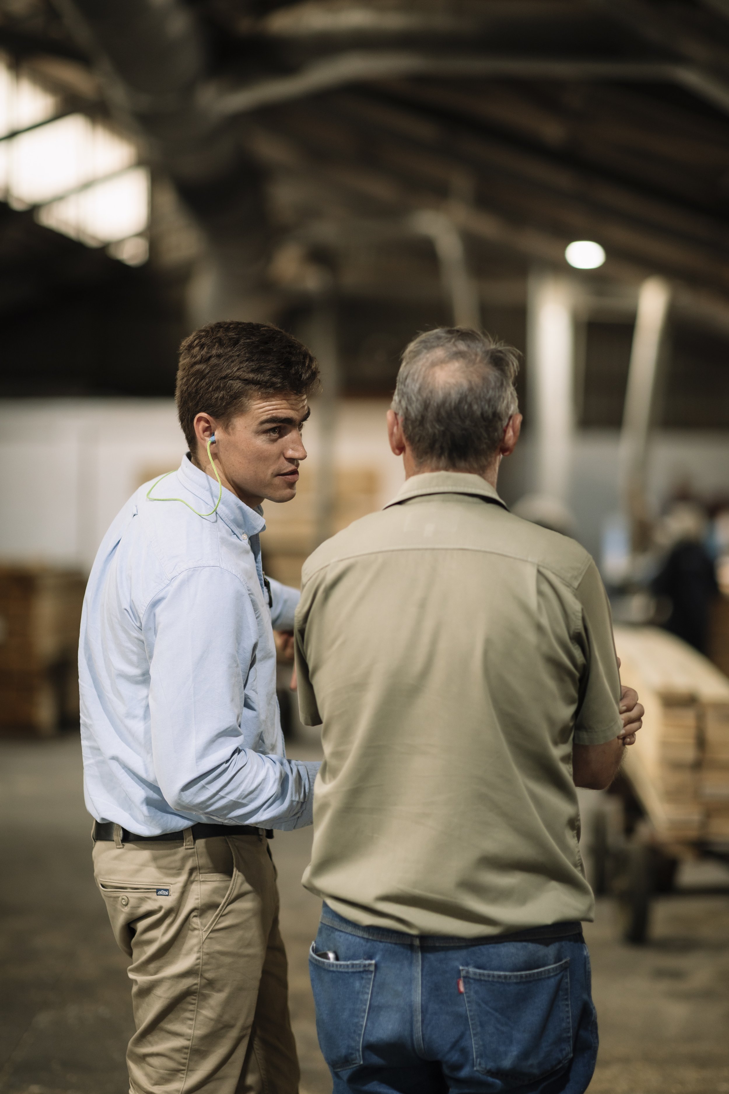 Two men are engaged in conversation in what appears to be a warehouse or industrial setting. One man is facing away from the camera, wearing a beige shirt and blue jeans. The other man is facing slightly to the side, wearing a light blue shirt with earphones in his ears.