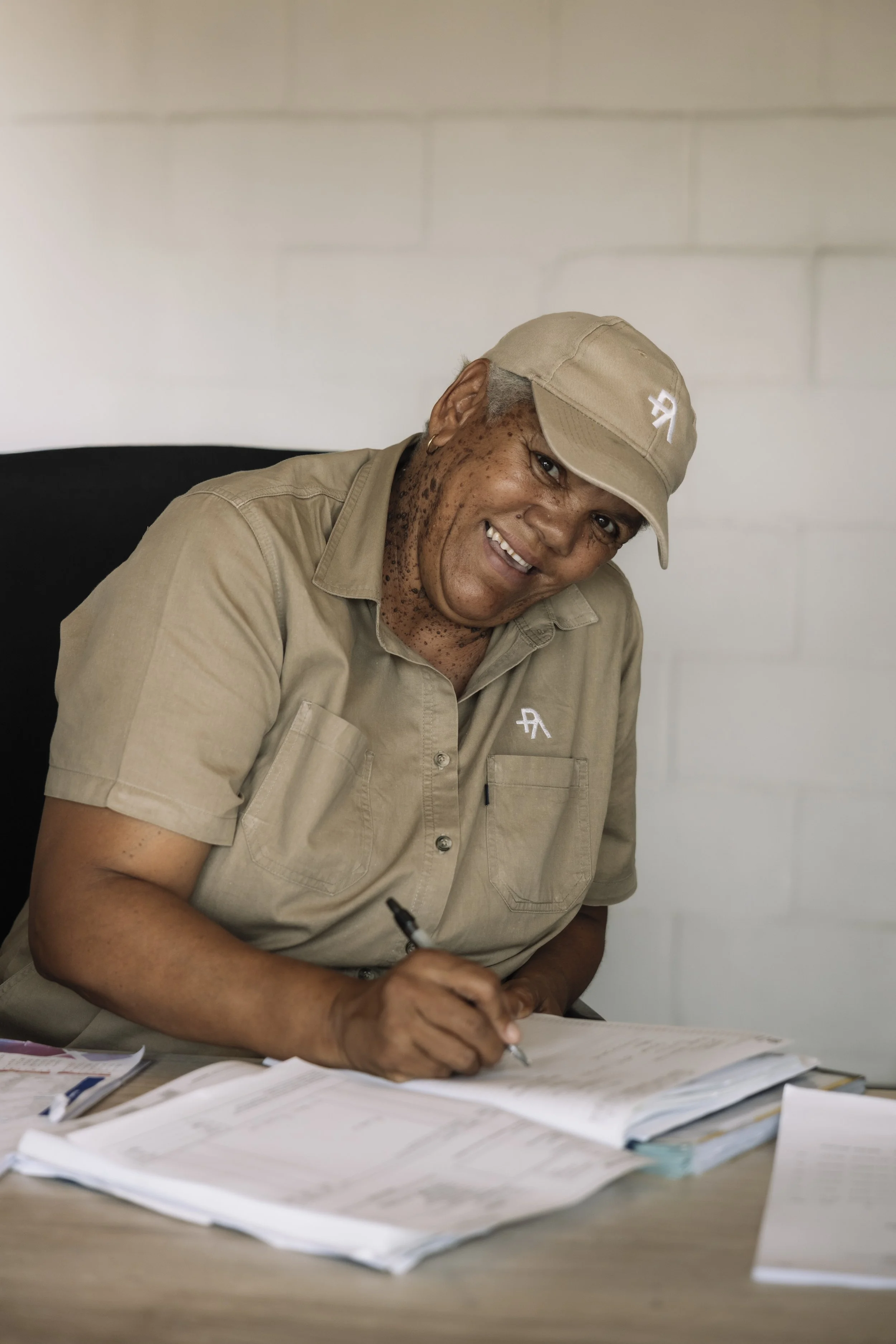A woman with short gray hair, wearing a beige cap and uniform, smiling and signing documents at a desk covered with papers.