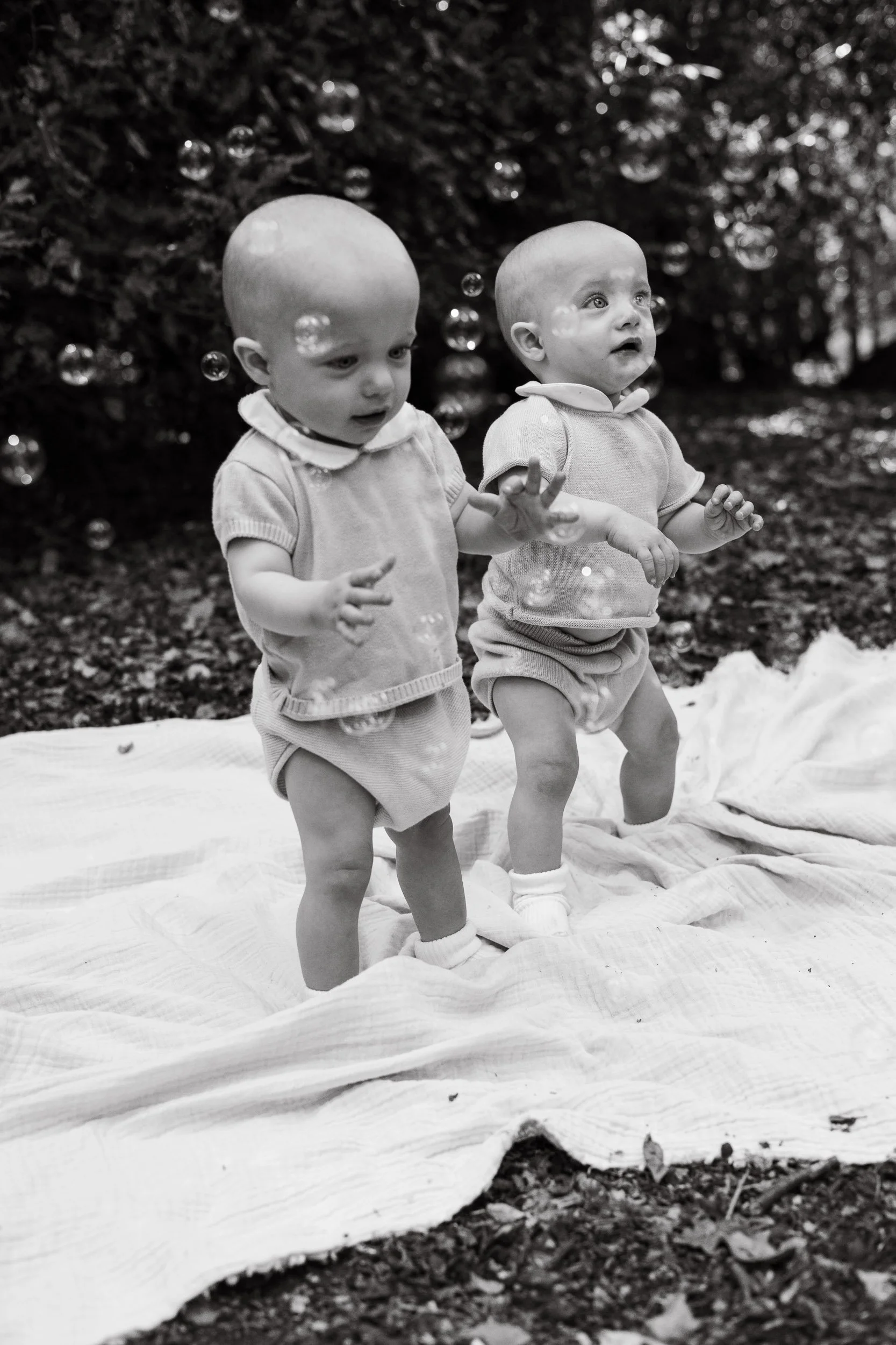 Twin babies standing on a white blanket catching bubbles during a natural light milestone photography session