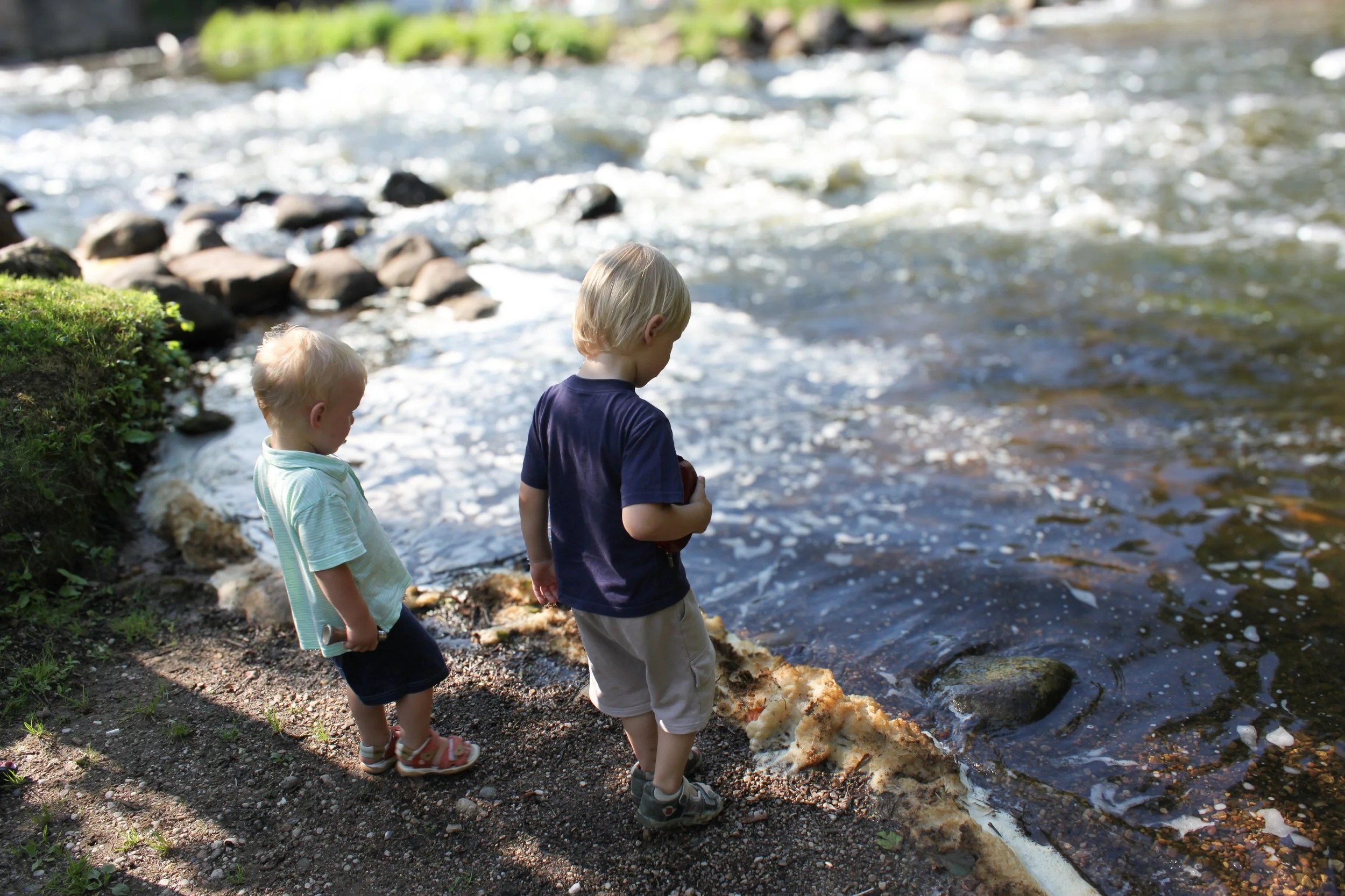 A bright and airy lifestyle portrait capturing a quiet moment of two brothers from behind, exploring a riverbank during an outdoor childhood session