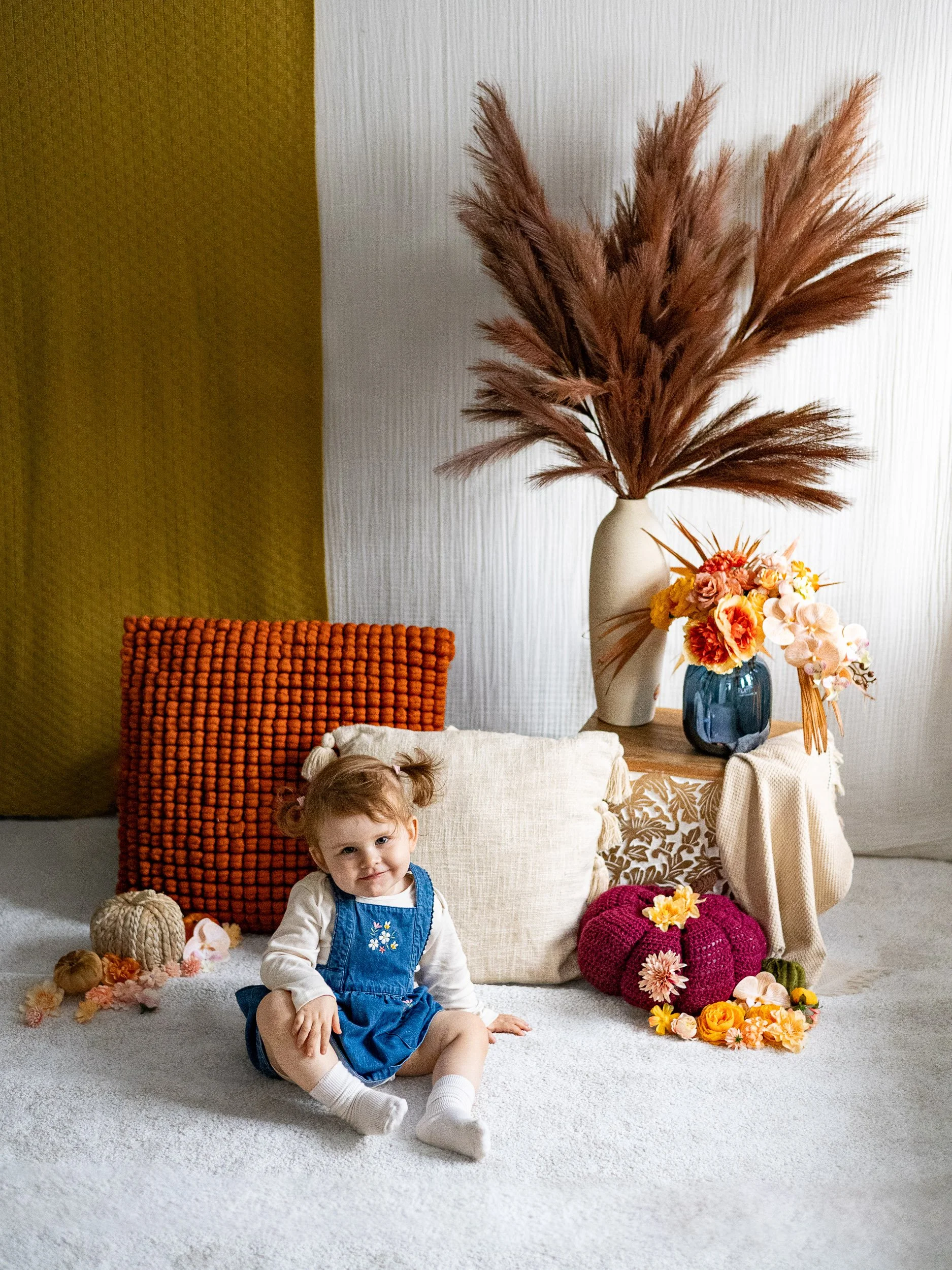 Toddler girl sitting in a denim overall dress on a white rug during an autumn family photography mini session in Surrey with Laura Rickute Photography.