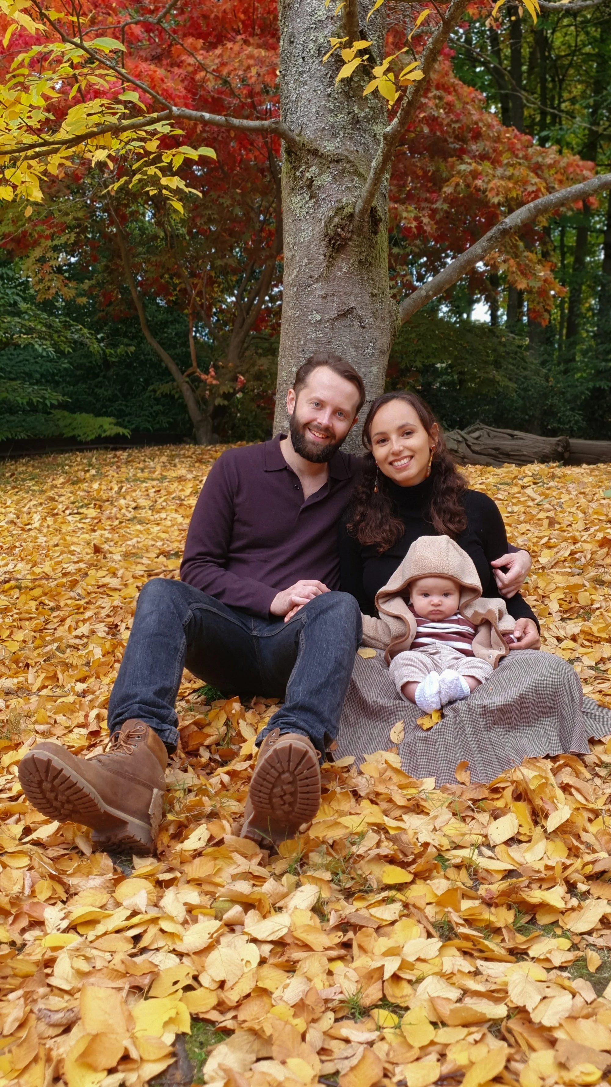 A heartwarming lifestyle portrait of a young family of two holding their baby under a tall tree in a beautiful autumn forest with golden leaves