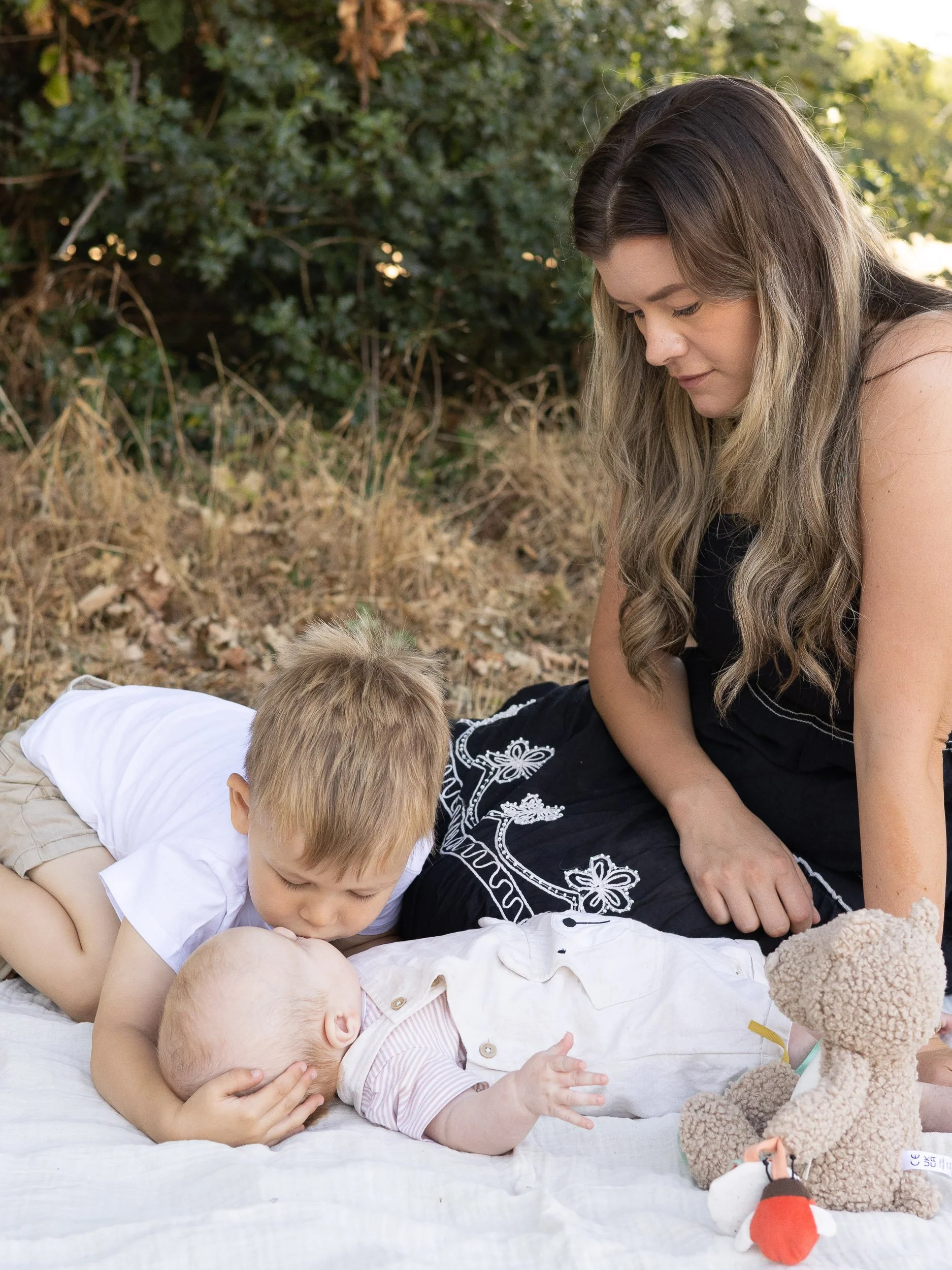 Mother sitting on a blanket outdoors in Surrey watching her young son gently kiss his newborn sibling's head during a natural documentary lifestyle photography session.