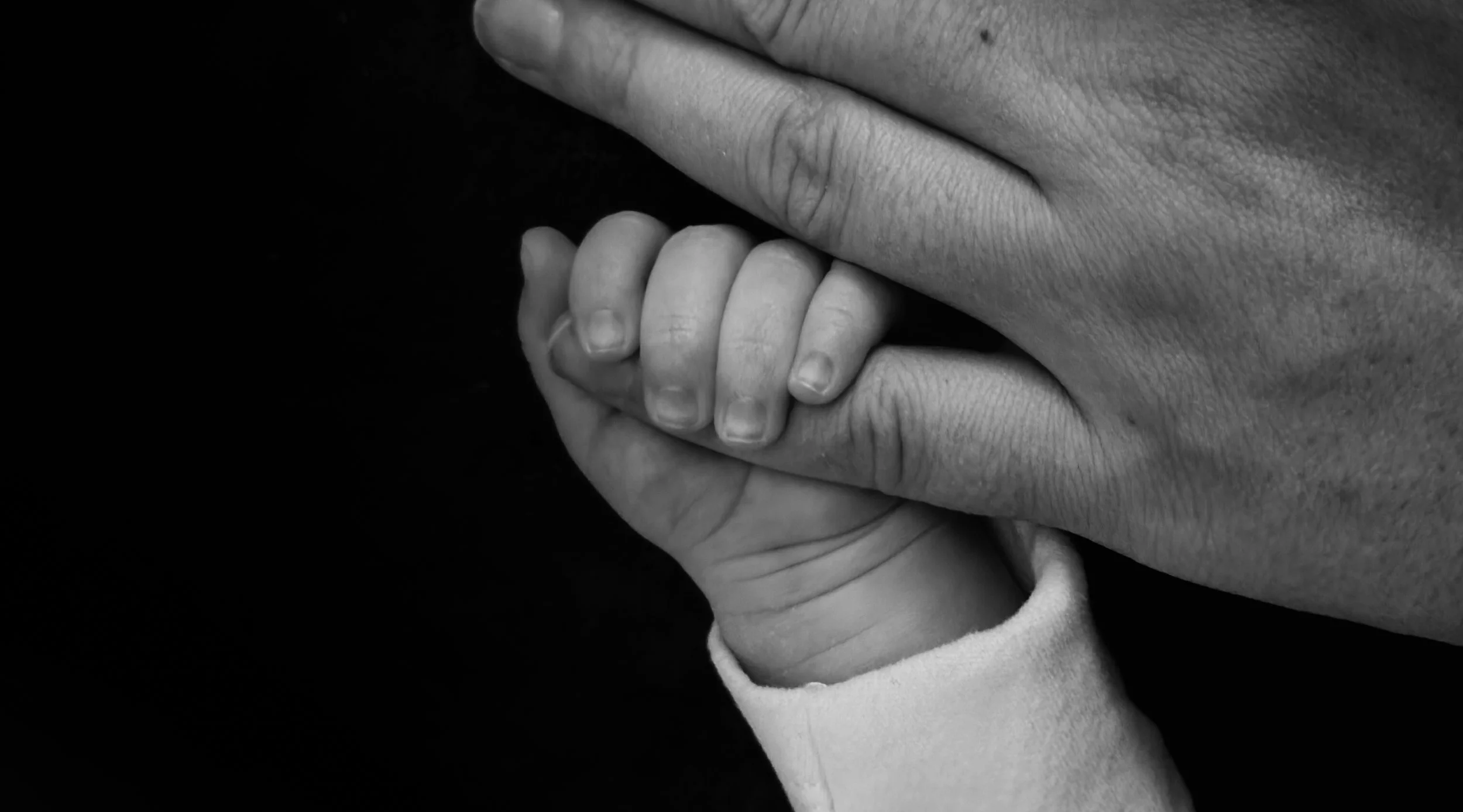 A close-up studio portrait capturing the tender connection of a tiny baby holding their mum's hand against a solid black background