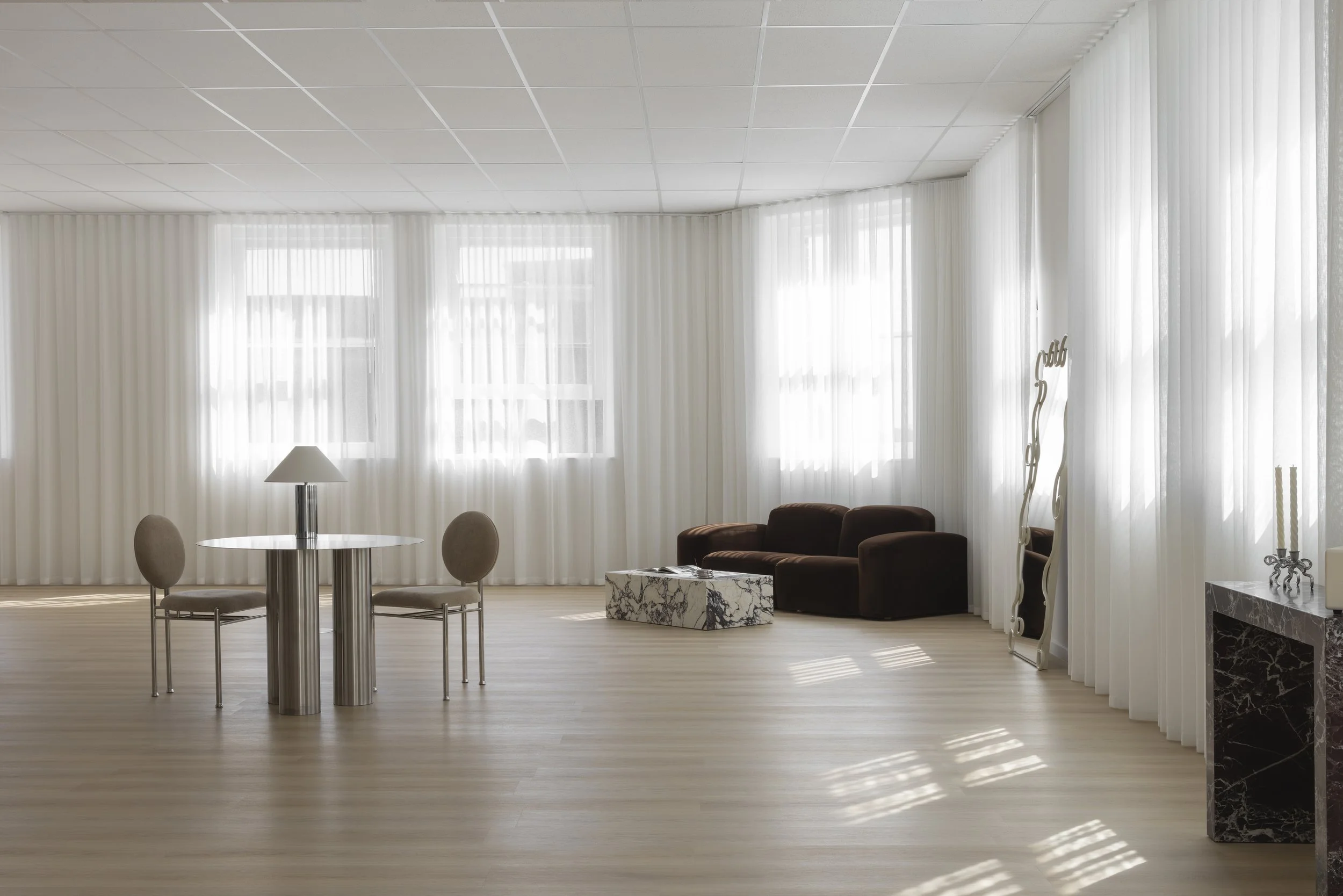Living room with large windows covered by white curtains, brown sofa, round table with beige chairs, floor lamp, marble fireplace, and decorative sculpture. Content, photography & event studio Newcastle NSW. 