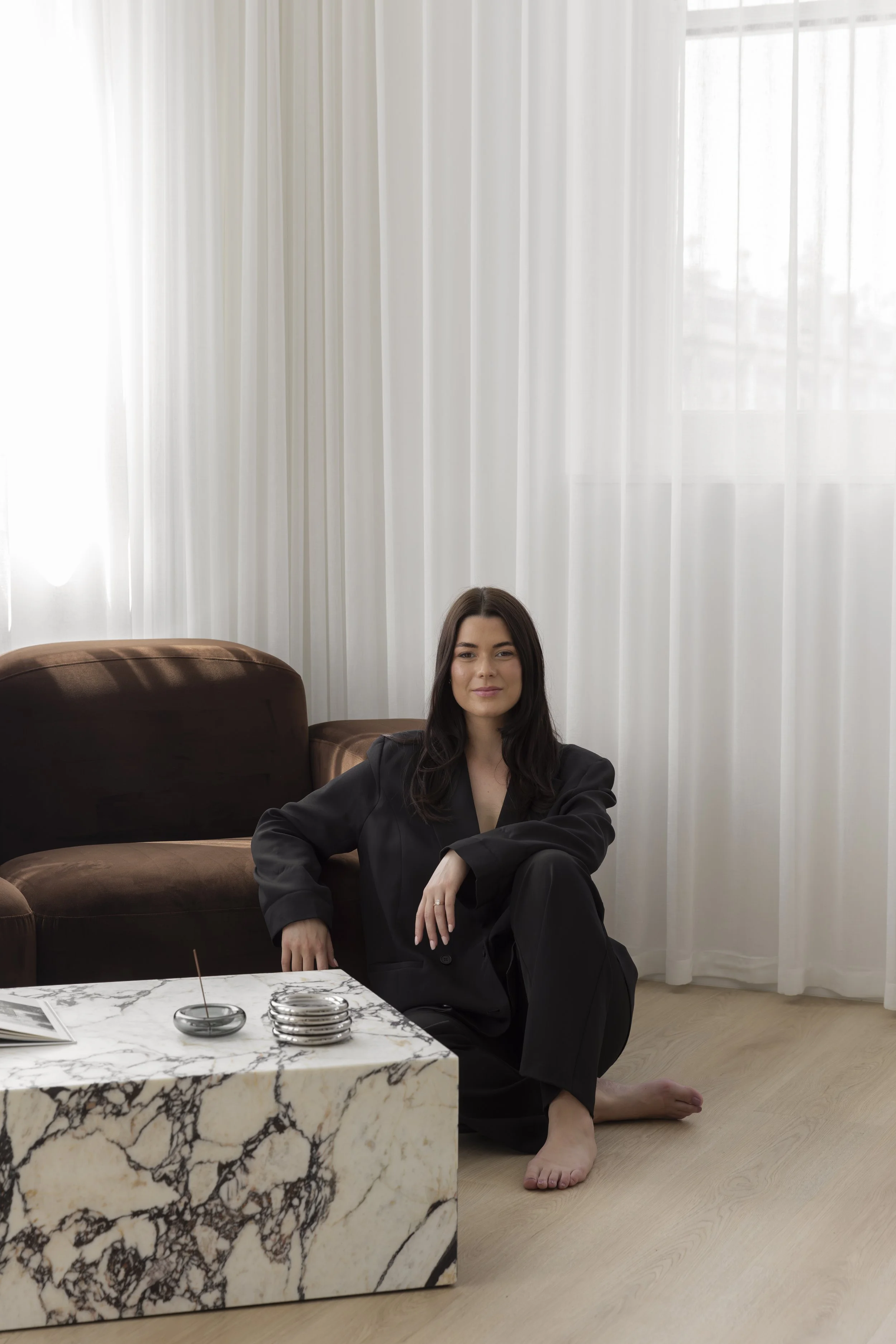 A woman with sitting on the floor in a black outfit, next to a marble table, in a room with light wooden flooring, sheer white curtains, and a brown couch in the background. Content, photography & event studio Newcastle NSW.