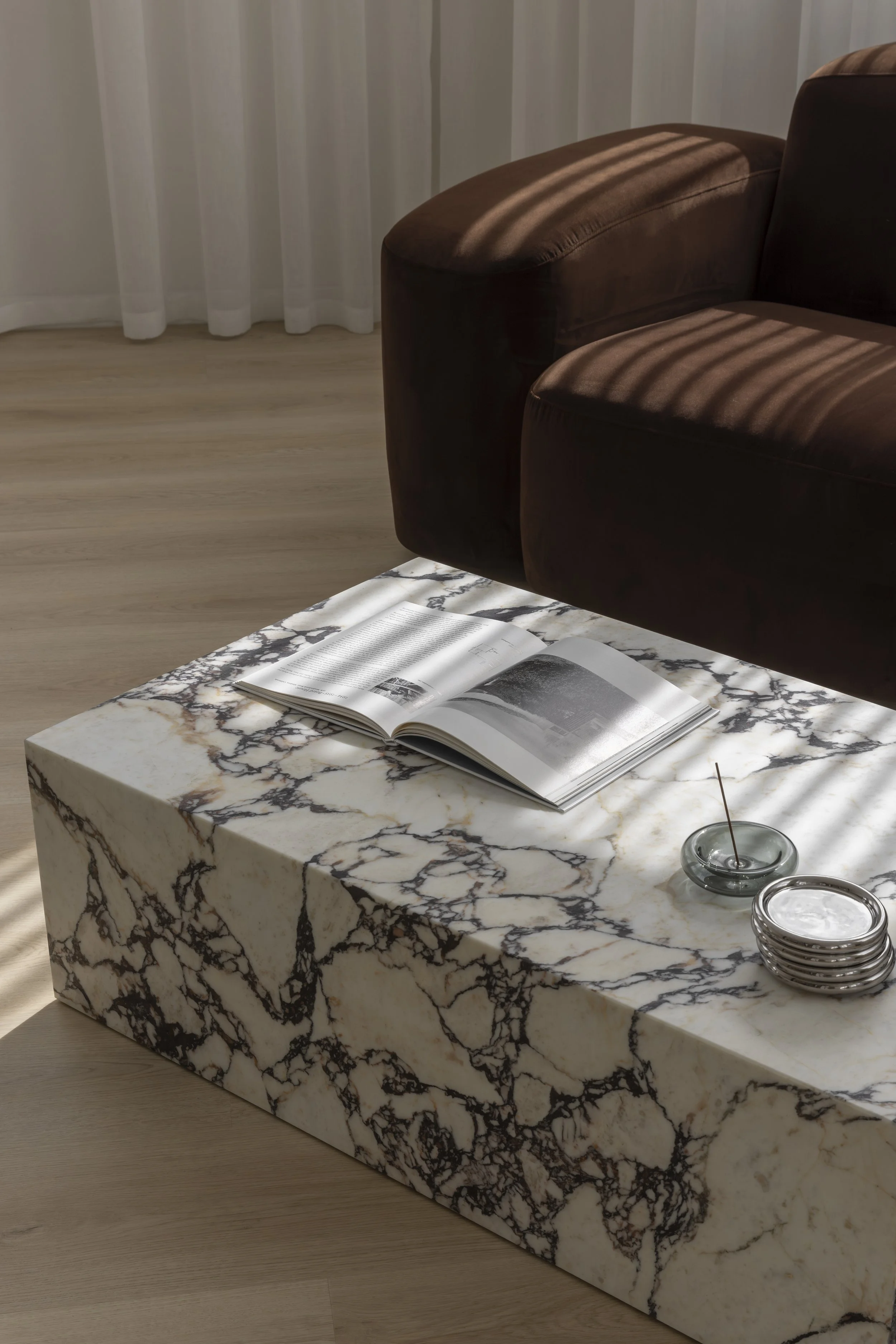 A marble coffee table with black and gold veining, topped with an open magazine, a small bowl with water and a stirring stick, and a glass container, sits beside a brown leather sofa in a sunlit living room with white curtains.