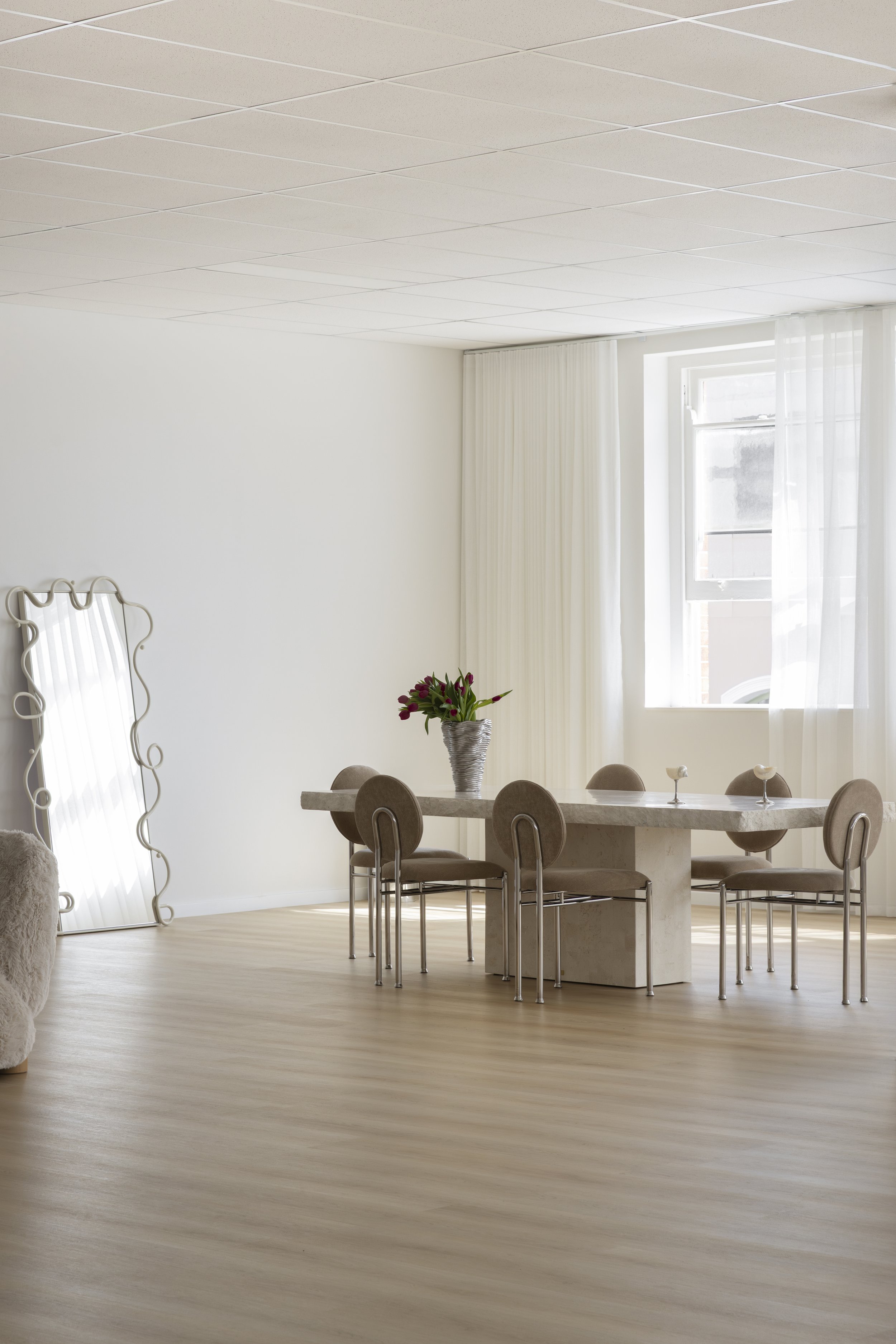 A bright dining room with a marble table surrounded by eight beige chairs, a vase with red flowers, large windows with sheer white curtains, a decorative mirror, and light wood flooring. Content, photography & event studio Newcastle NSW.