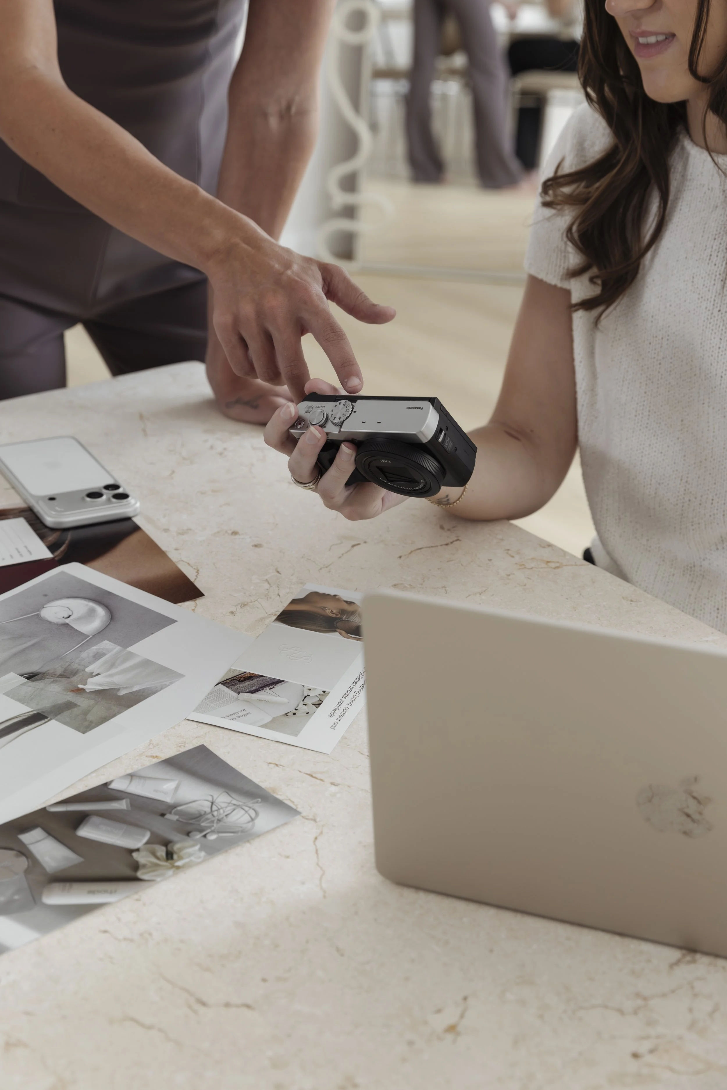 A woman holding a camera and another person pointing at it during a discussion at a desk with printed photos, laptops, and papers. Content, photography & event studio Newcastle NSW.
