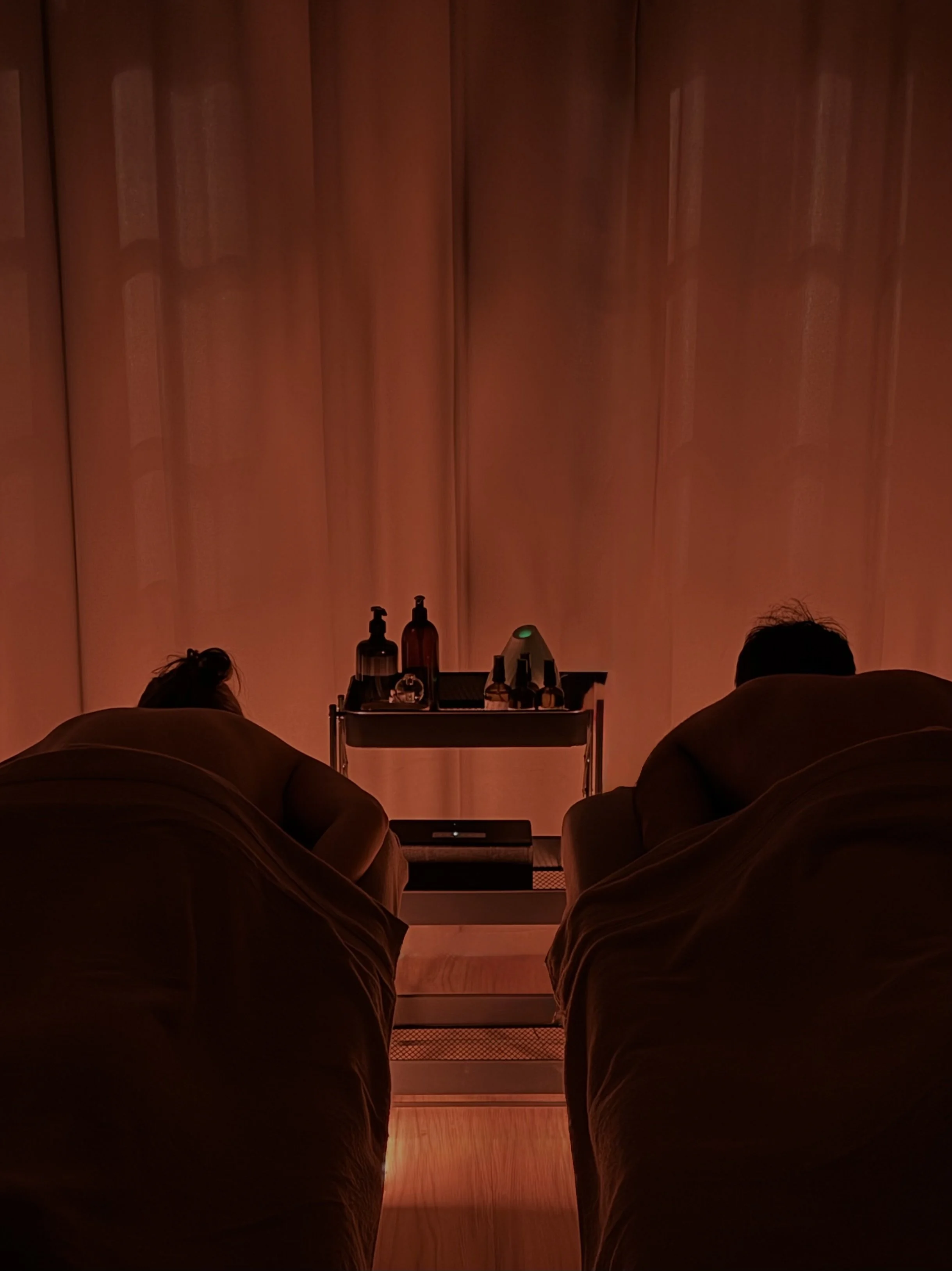A couple lying on the massage table, receiving a massage in a dimly lit spa room, facing a curtain with bottles on a small table in front.