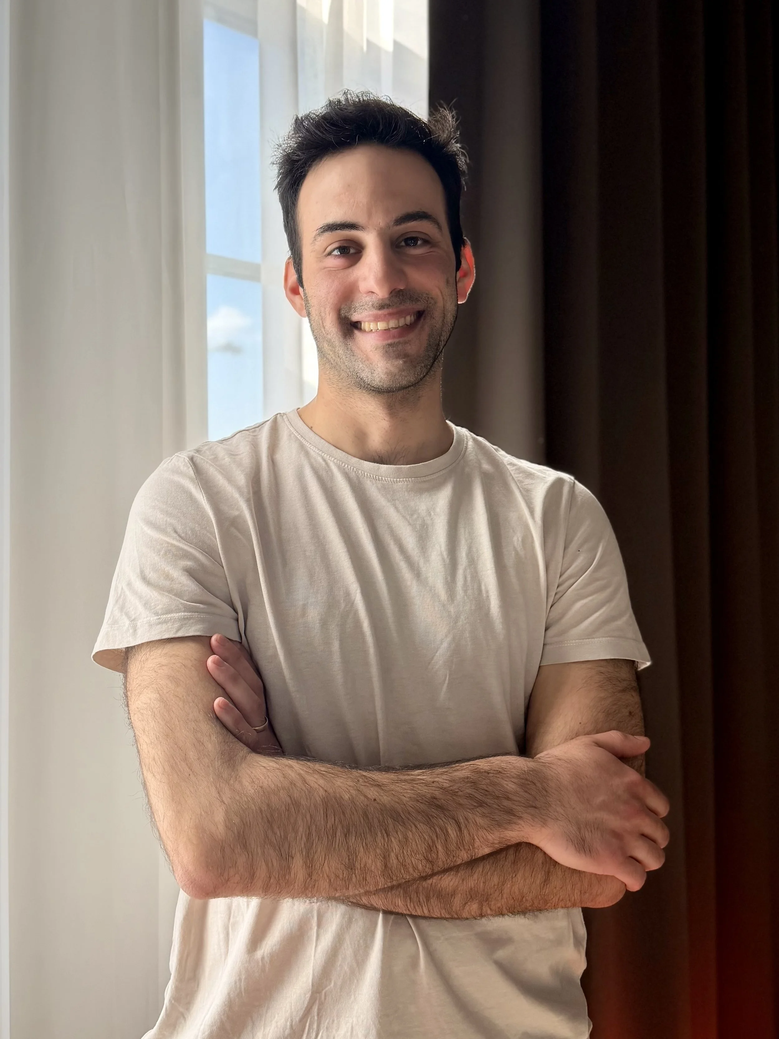 A young man with dark hair, light skin, and facial stubble smiling with arms crossed, standing in front of a window with white light and curtains.