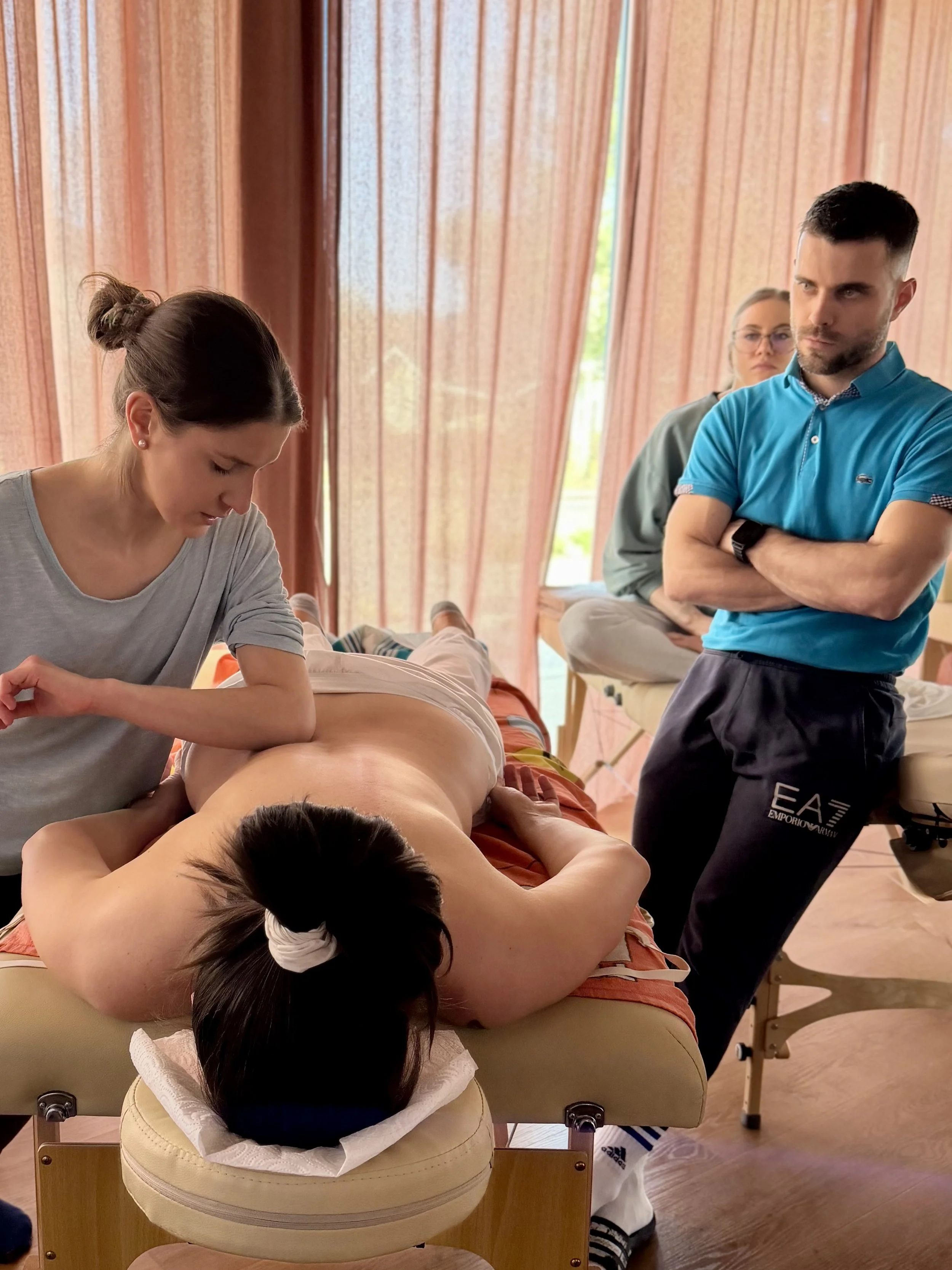 A woman is receiving a massage on her back while lying on a massage table in a room with pink curtains. Two people, a woman and a man, are standing nearby watching. The man is wearing a blue shirt with crossed arms, and the woman is behind him wearin