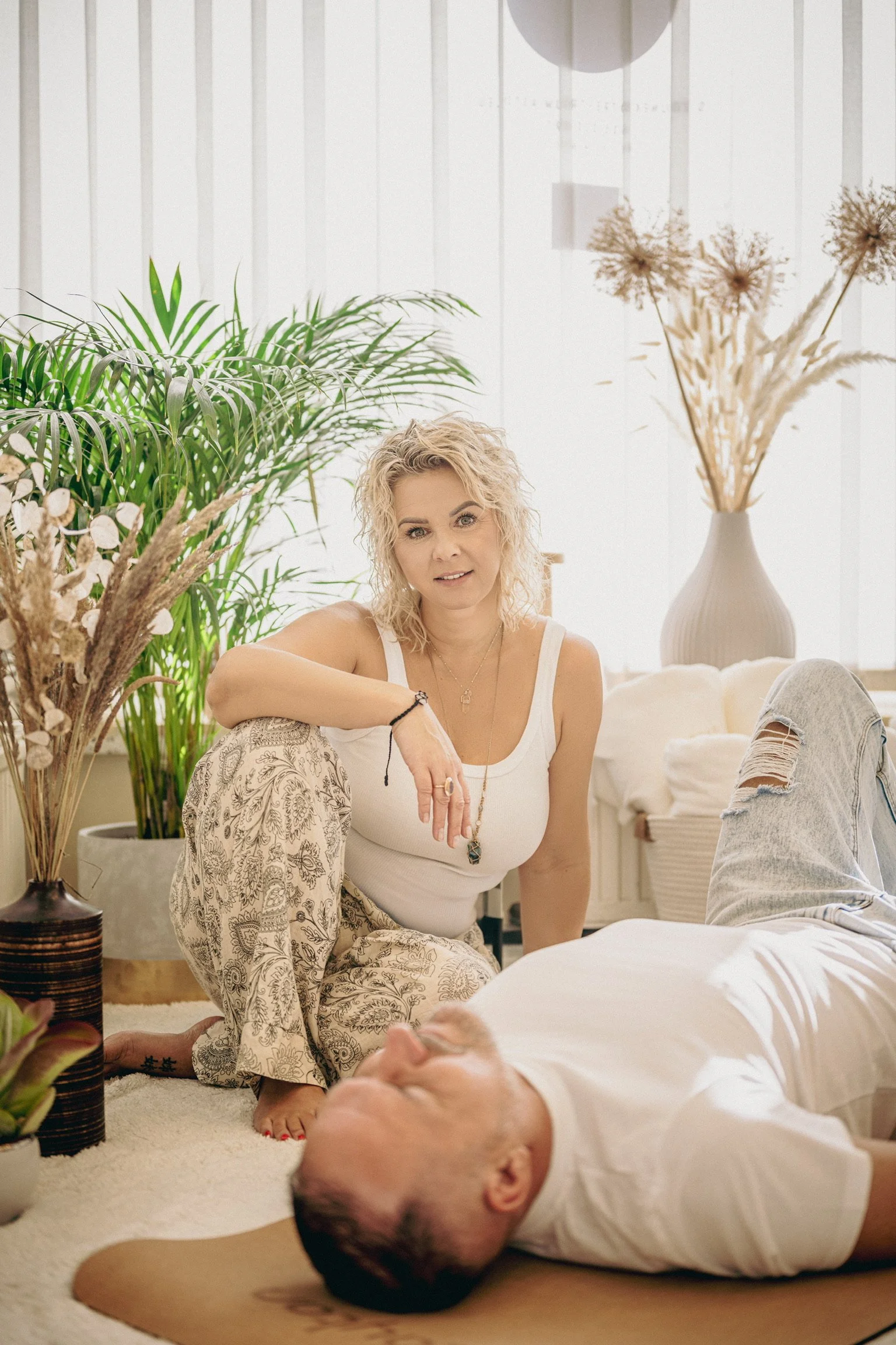 A woman with curly blonde hair, wearing a white tank top and patterned pants, sitting on the floor, looking at a man lying down in front of her. The setting is a bright, cozy room with large plants and decorative dried flowers in vases.