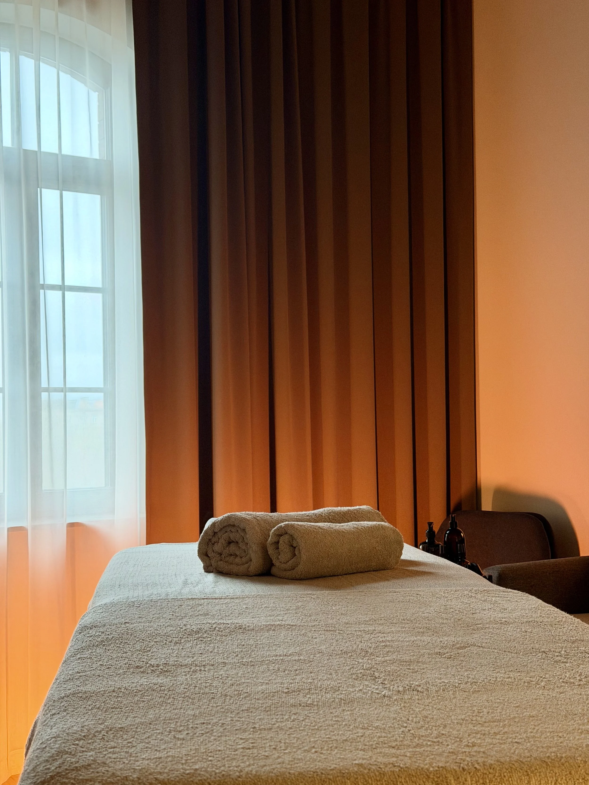 A cozy bedroom with a bed covered in a textured beige blanket, two neatly rolled towels on top, a window with sheer curtains letting in natural light, and a wooden wall or partition behind the bed, with bottles and a chair on the side.