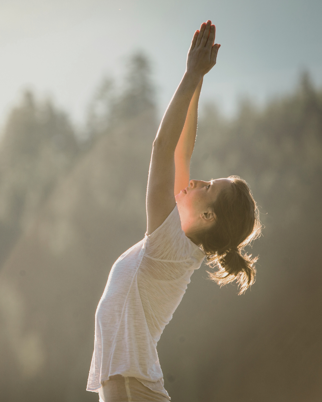 A young woman in athletic clothing stretching outdoors at sunrise or sunset, with trees in the background.