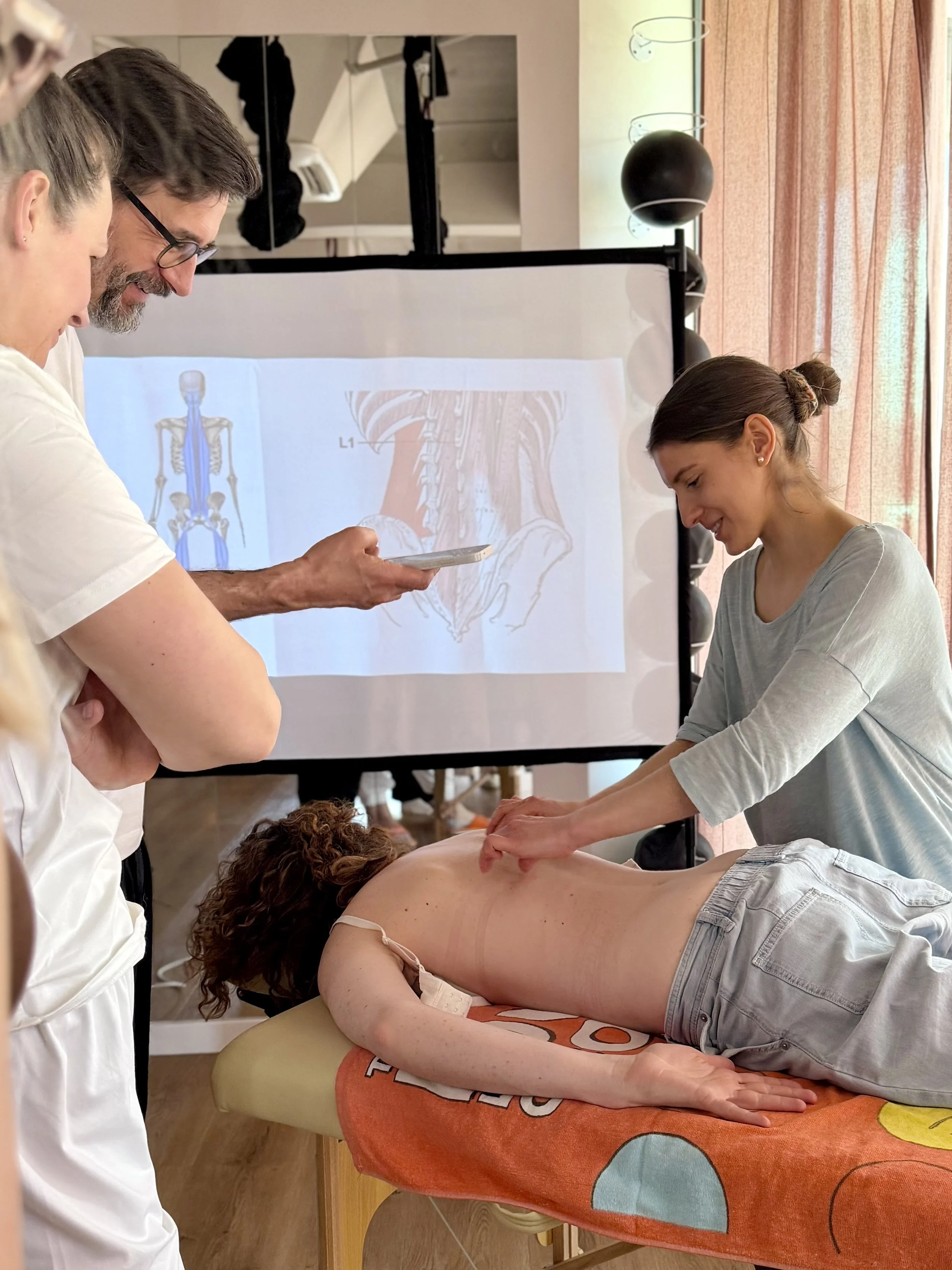 A person giving a back massage to a woman lying face down on a massage table, with two people observing beside them and a doctor or instructor standing nearby. A presentation slide with anatomical diagrams is projected on a screen in the background.