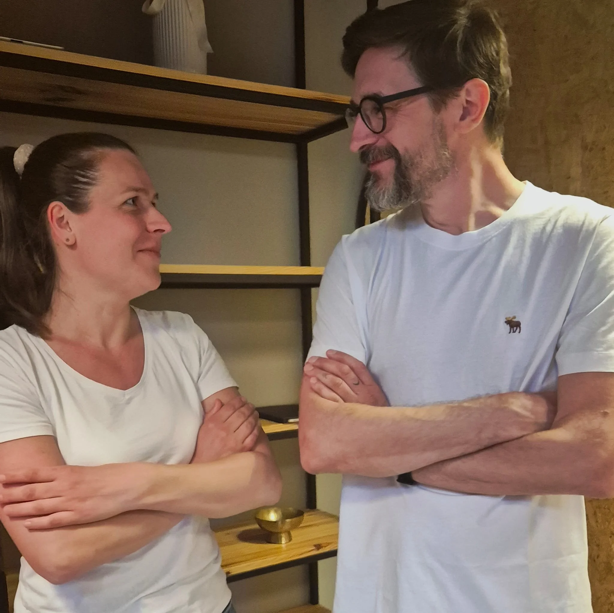 A woman and a man standing closely, facing each other with arms crossed, gazing into each other's eyes in a warmly lit room. The woman has brown hair tied back and is wearing a white shirt. The man has glasses, a beard, and is wearing a white T-shirt with a small animal graphic on the lower corner.