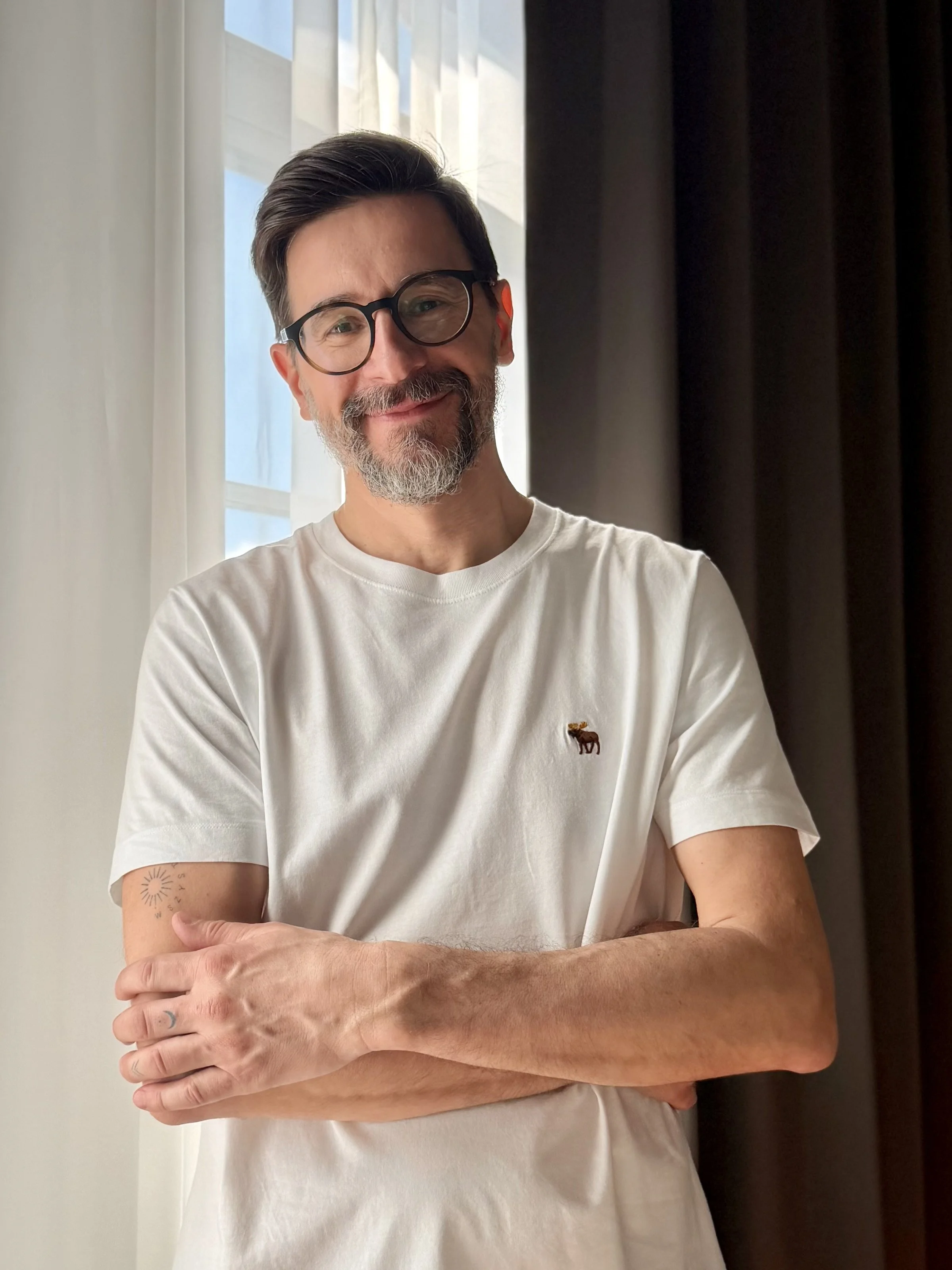 A man with a beard and glasses, smiling, wearing a white t-shirt with a small embroidered animal logo, standing against a background of a window with white curtains and dark drapes.