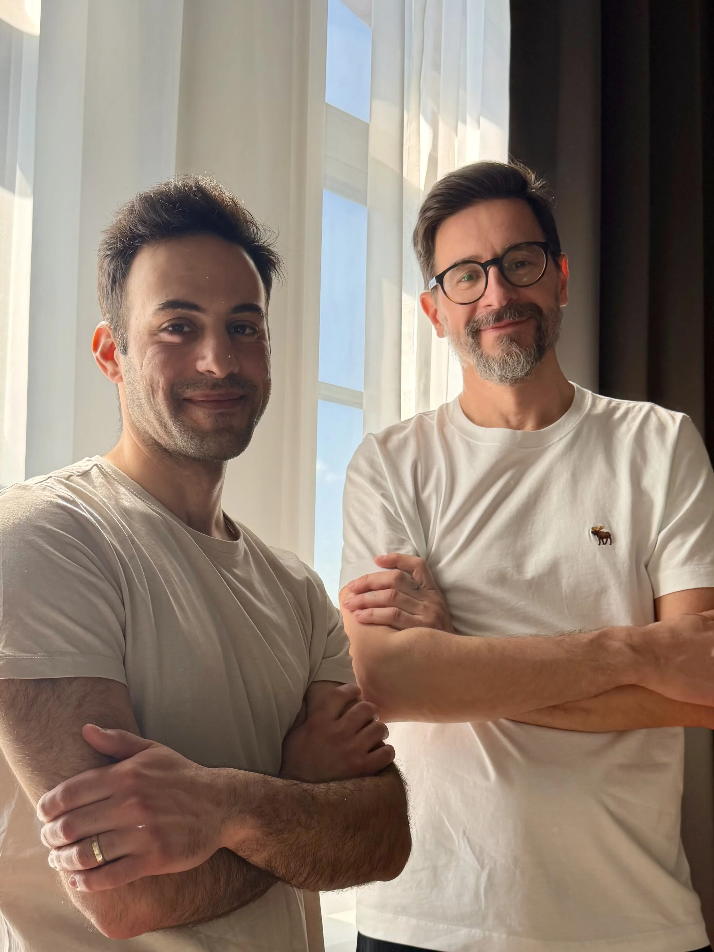 Two men standing together inside near a window with white curtains. One man has short dark hair, a slight beard, and is wearing a beige t-shirt. The other man has short hair, glasses, and a beard, and is wearing a white t-shirt with a small logo of a