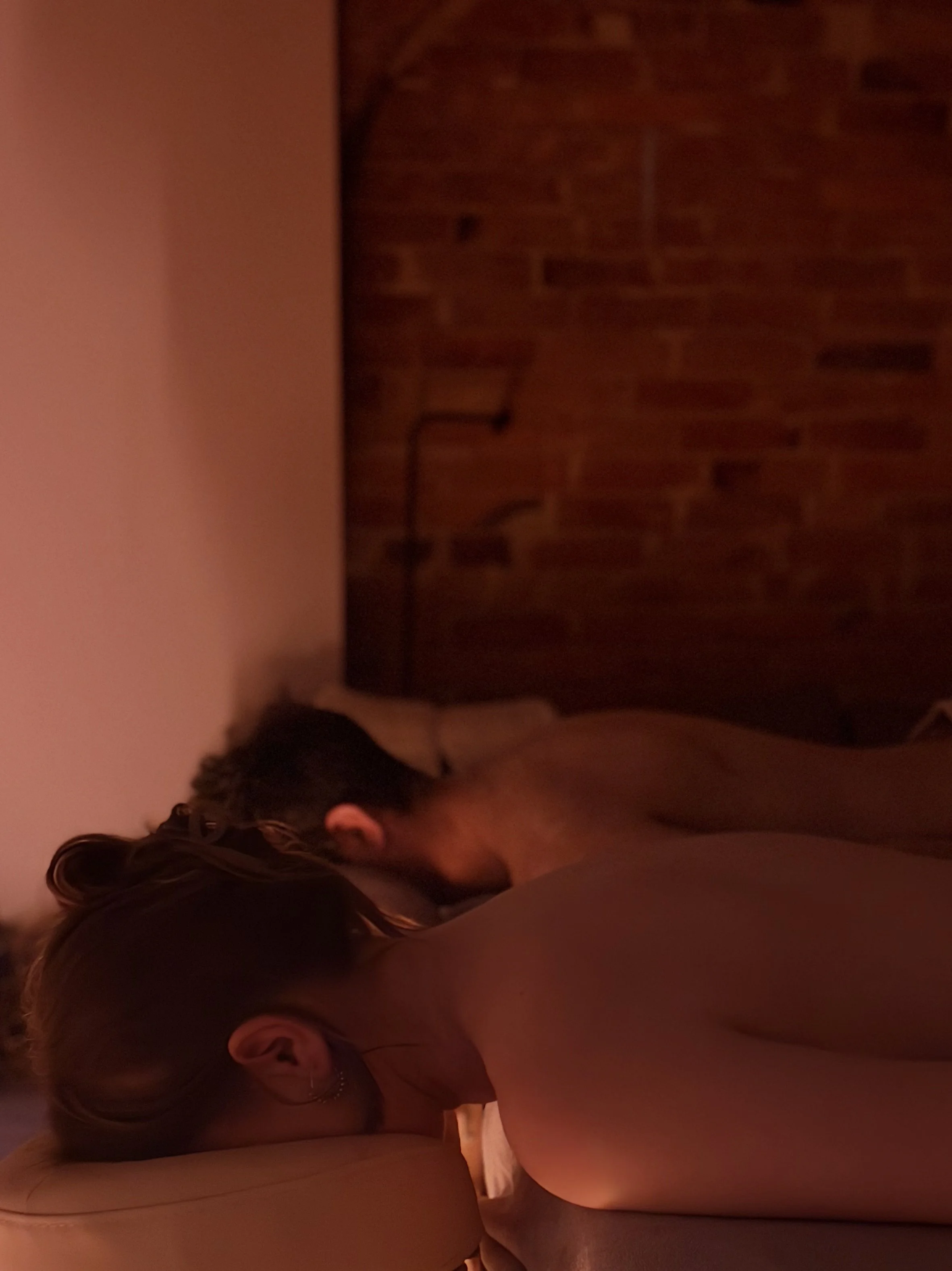 A couple lying on the massage table, receiving a massage in a dimly lit spa room, facing a curtain with bottles on a small table in front.