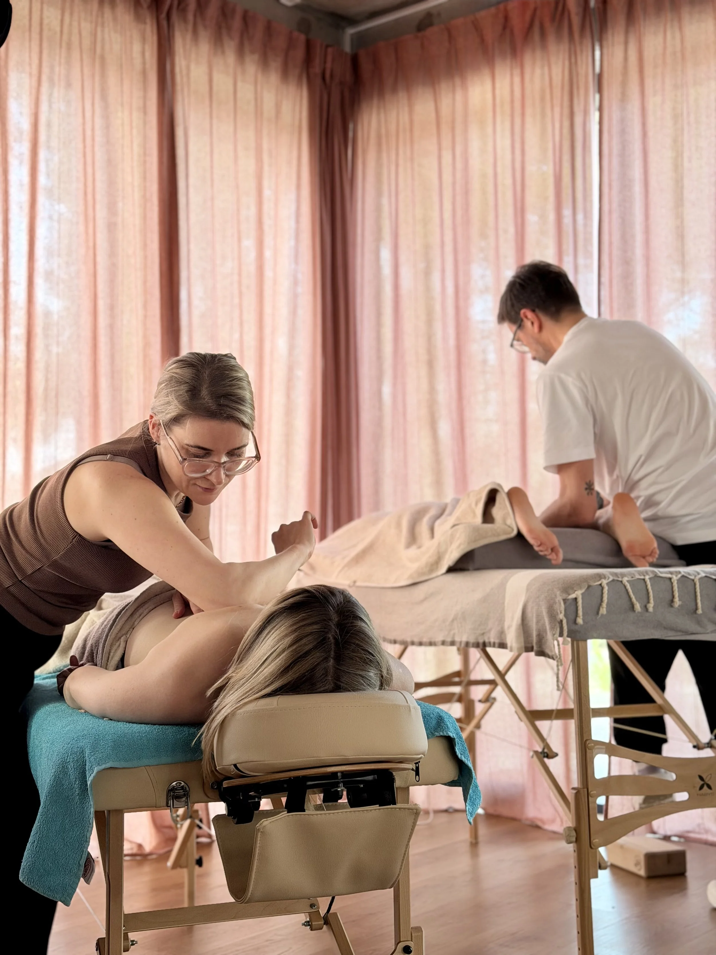 A woman lying face down on a massage table receiving a massage from a therapist, with a second therapist working on a person's back on a higher massage table nearby, in a room with pink curtains.