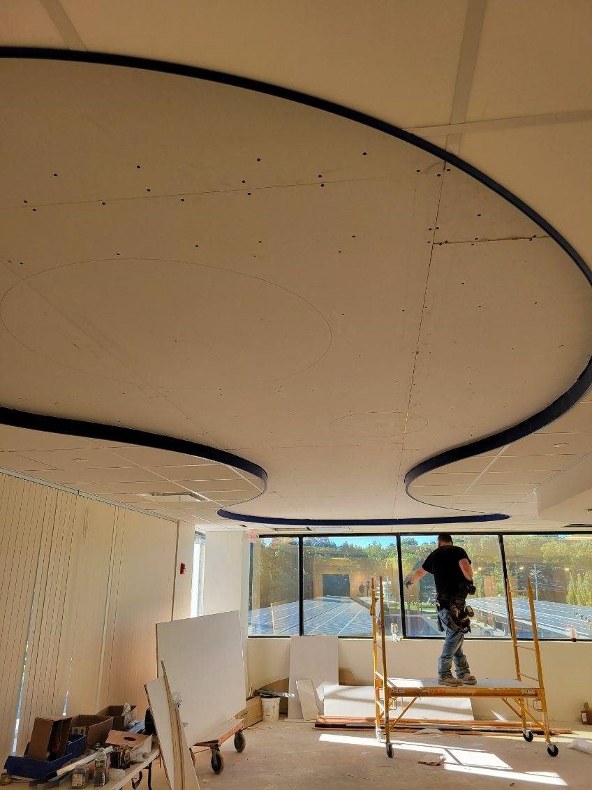 Construction worker standing on a scaffold installing or working on a ceiling with curved black accents, in a room with large windows and construction materials around.