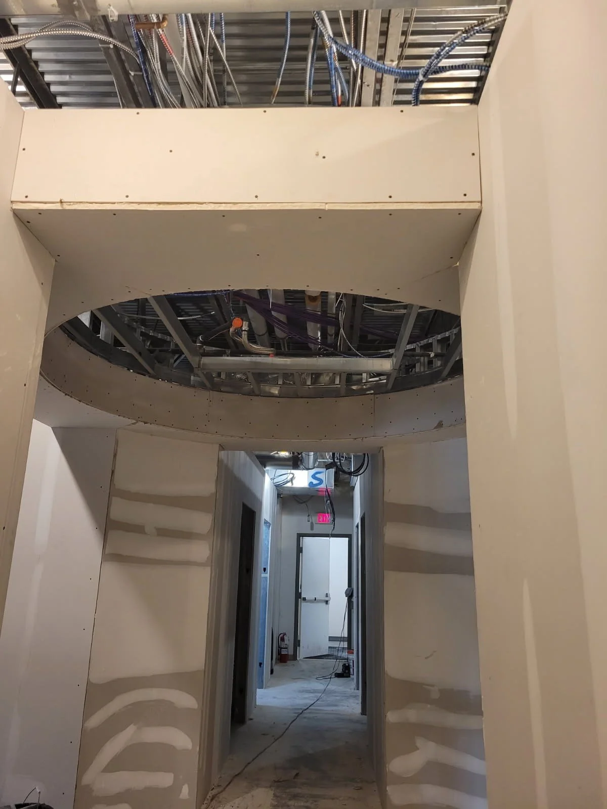 Construction site interior showing drywall walls, ceiling framing, and open ceiling with electrical wiring and metal ductwork.