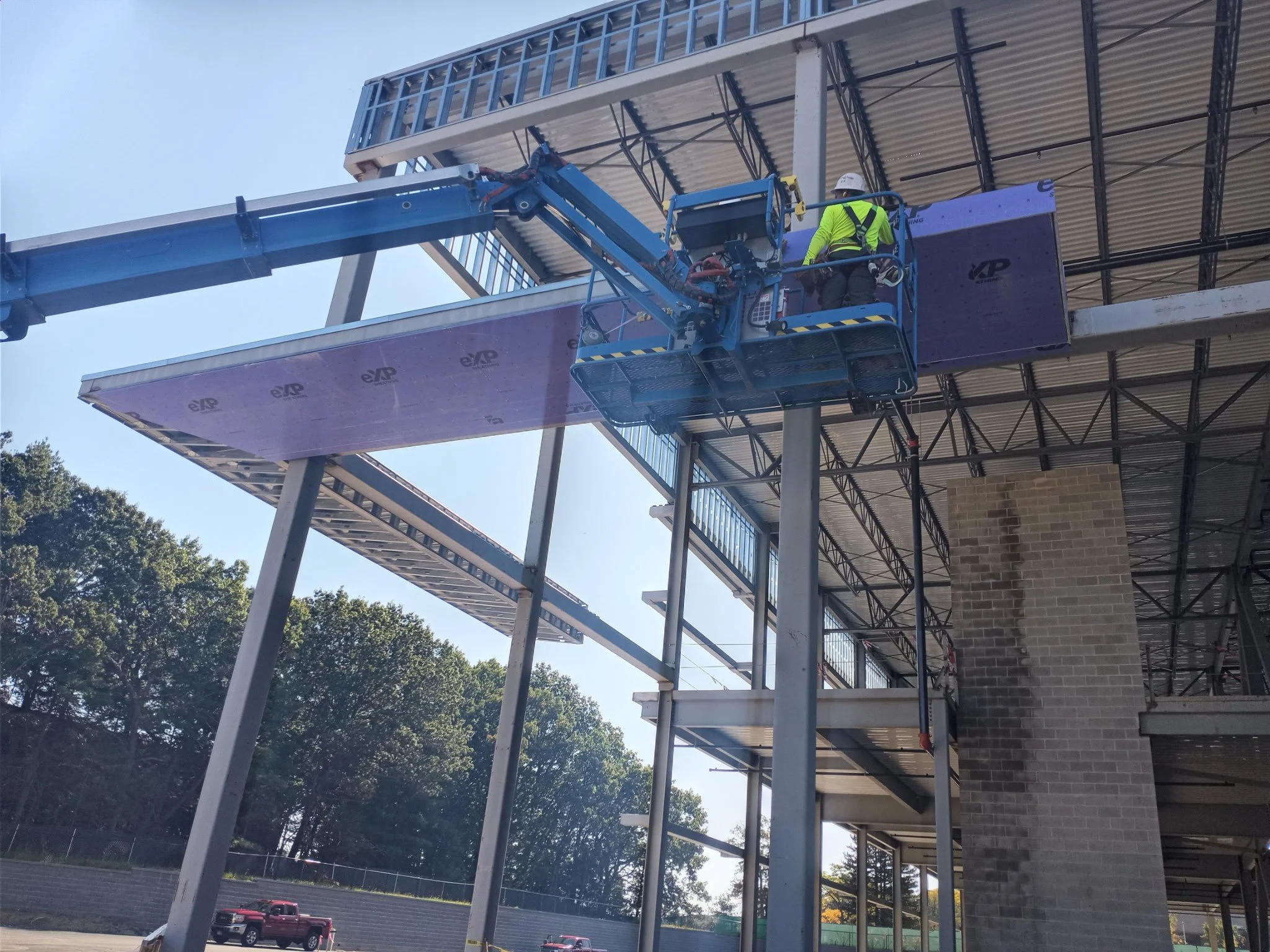 Construction worker in a yellow safety vest and white helmet operating a blue aerial lift near a building with brick walls and metal framework, against a clear blue sky with trees in the background.
