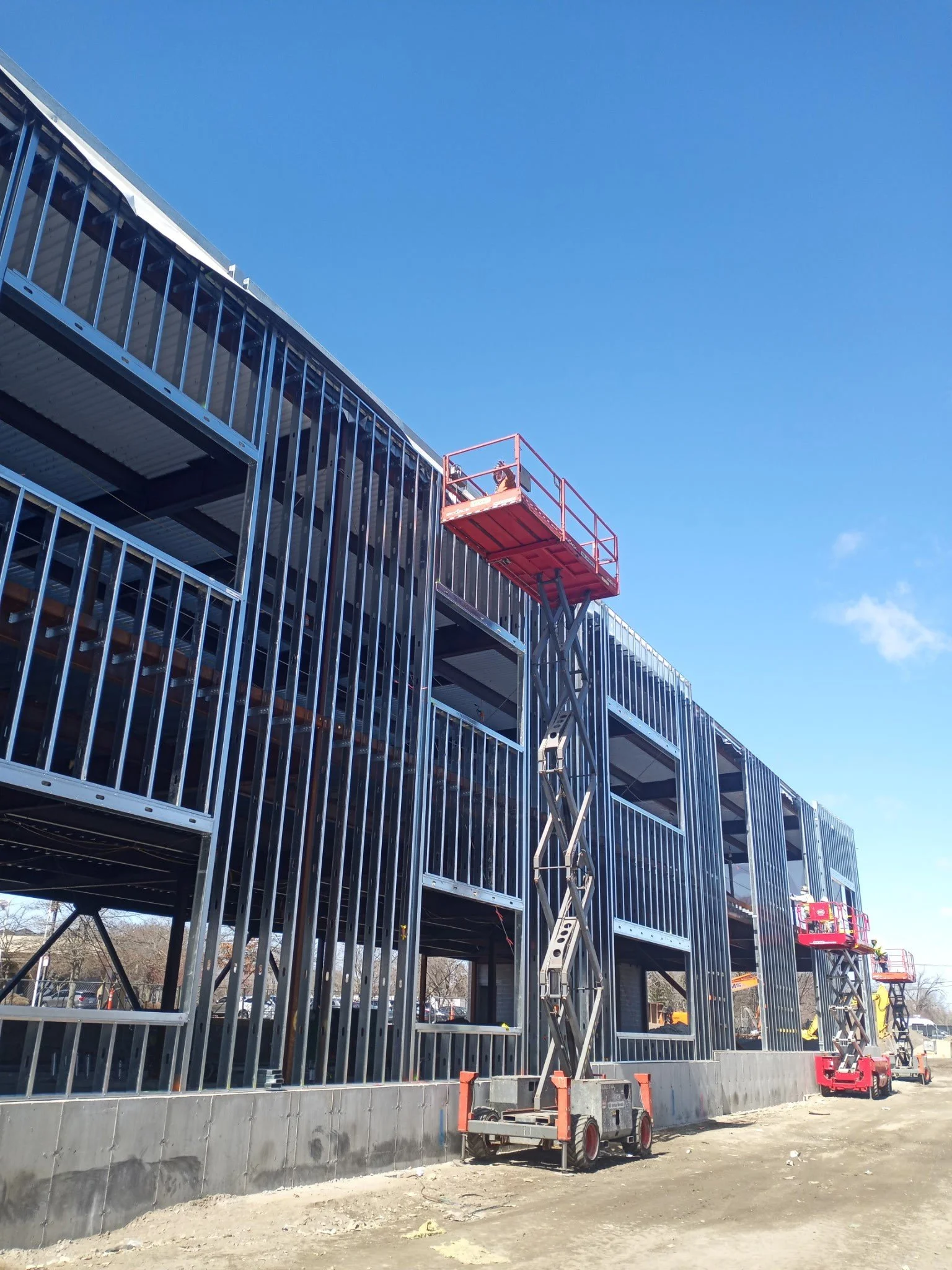 Construction site with a partially built multi-story building with metal framing, a red lift platform with a worker, and a concrete foundation at the base under a clear blue sky.