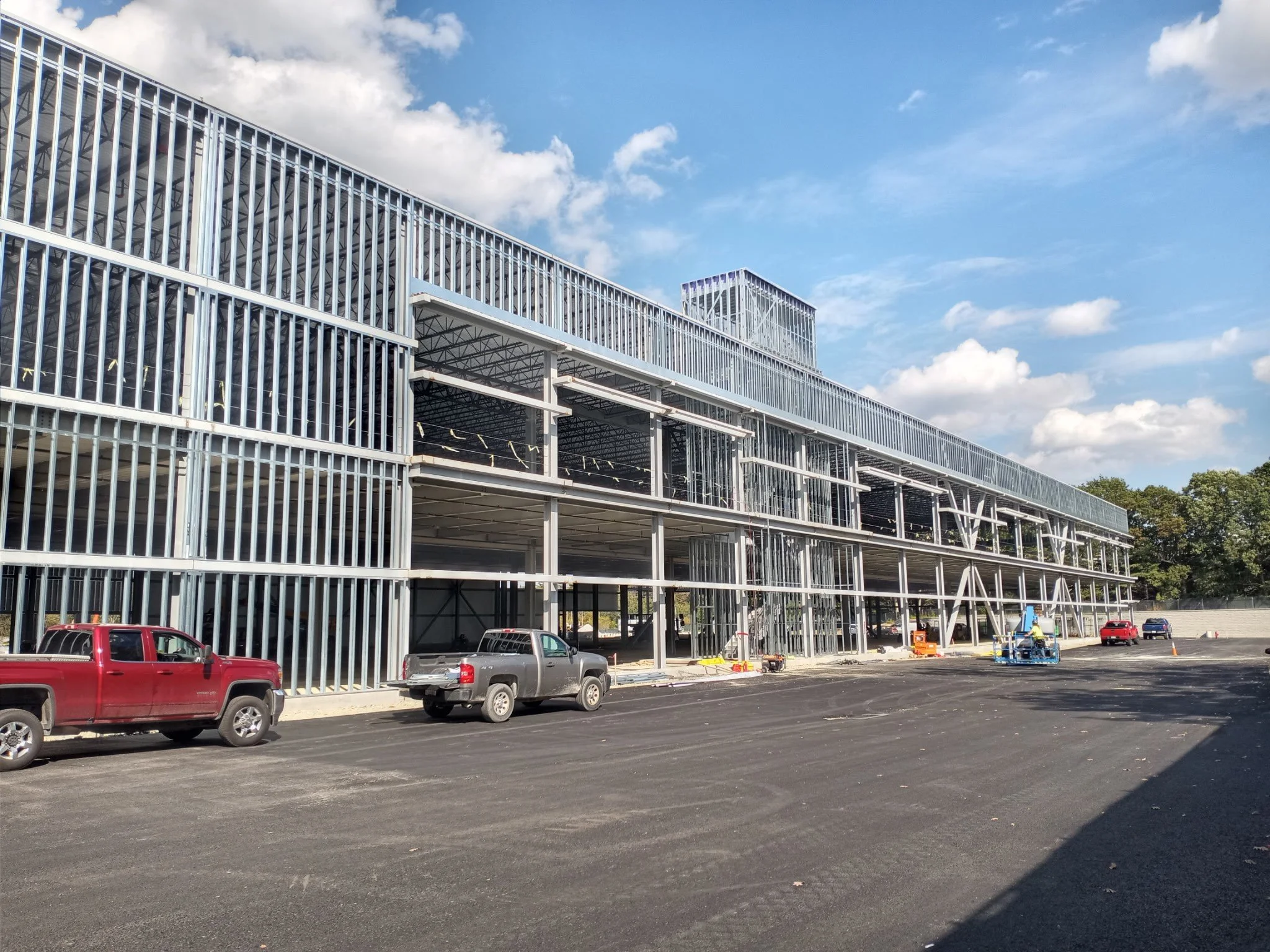 Exterior of a building under construction with steel framework, parked cars in the foreground, and a partly cloudy sky.