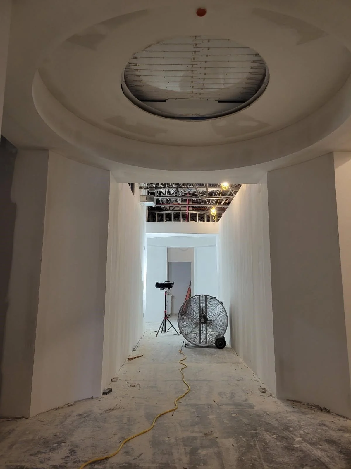 Construction hallway with unfinished ceilings, construction equipment including a large fan, tripod, and extension cord, and white walls in progress.