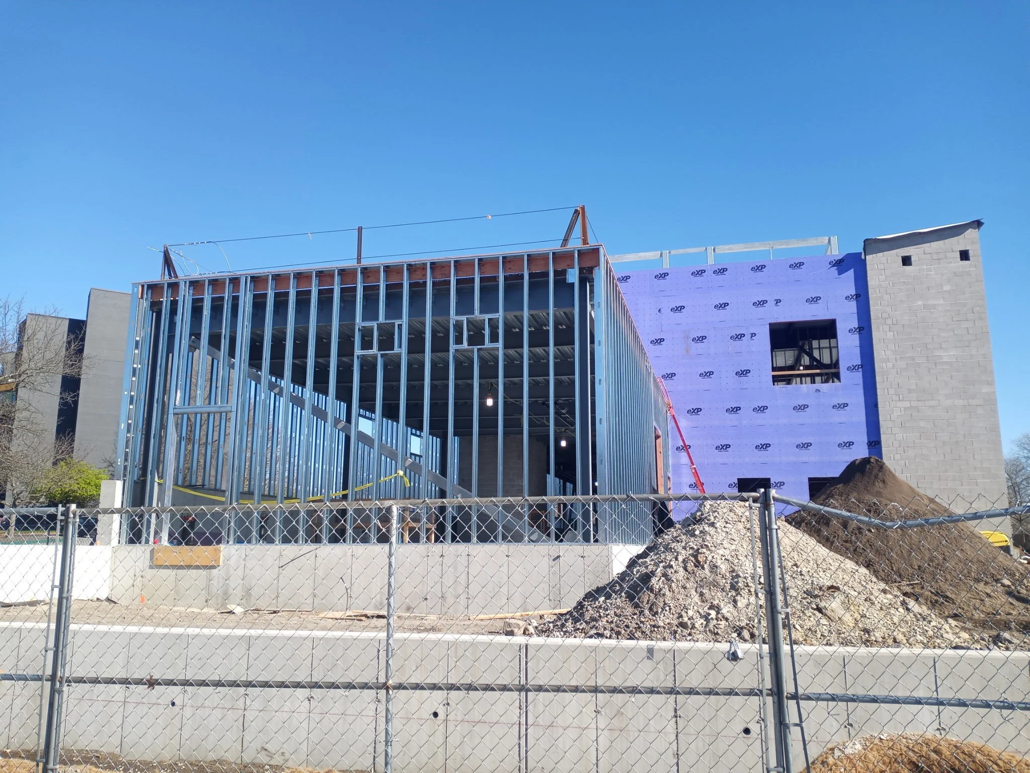 Building under construction with steel framework and partially installed exterior walls, surrounded by a chain-link fence and construction debris, against a clear blue sky.