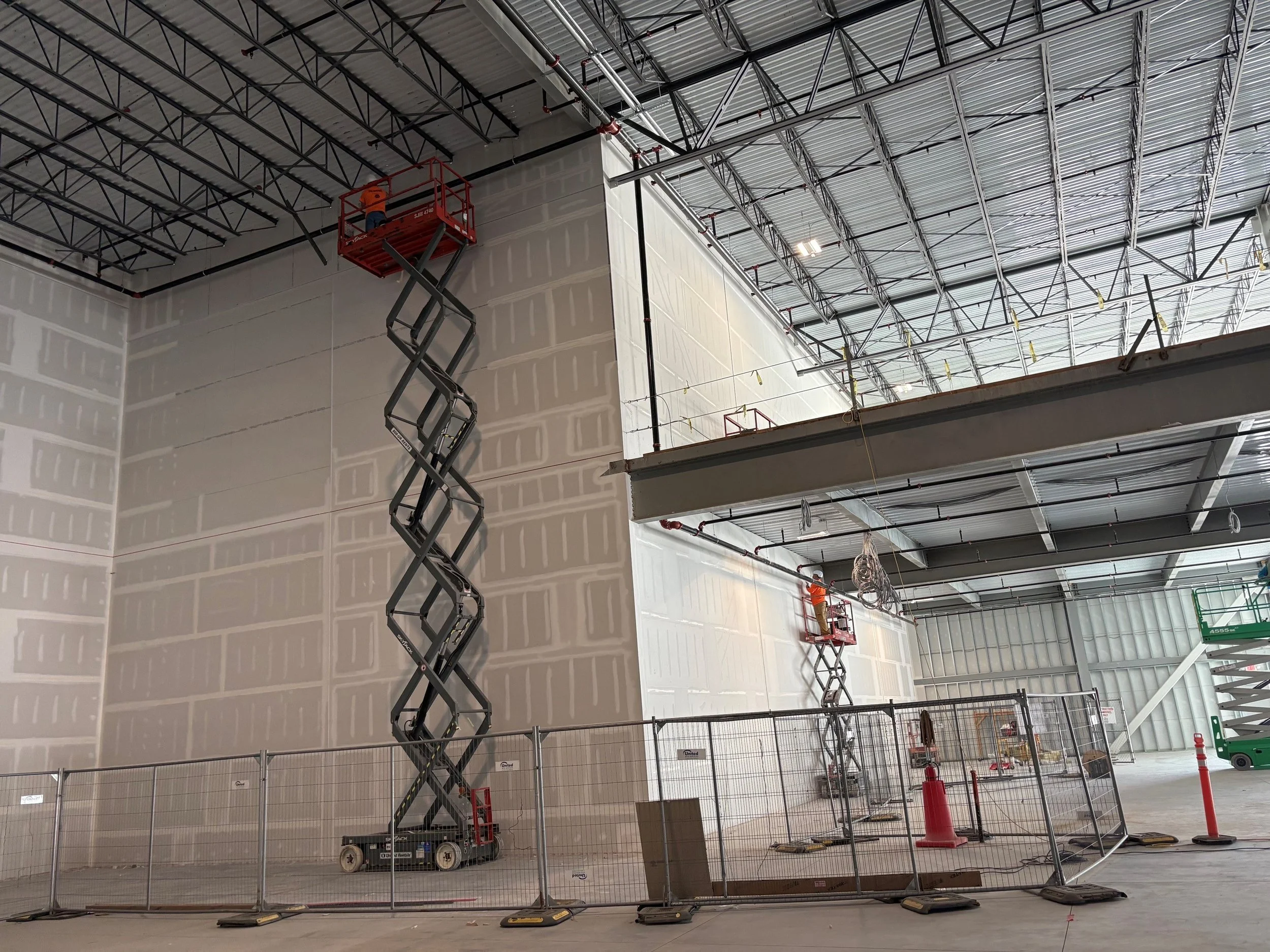 Construction workers on elevated platforms working on the ceiling of a large building with exposed metal framework and partially installed walls, surrounded by safety barriers and equipment.